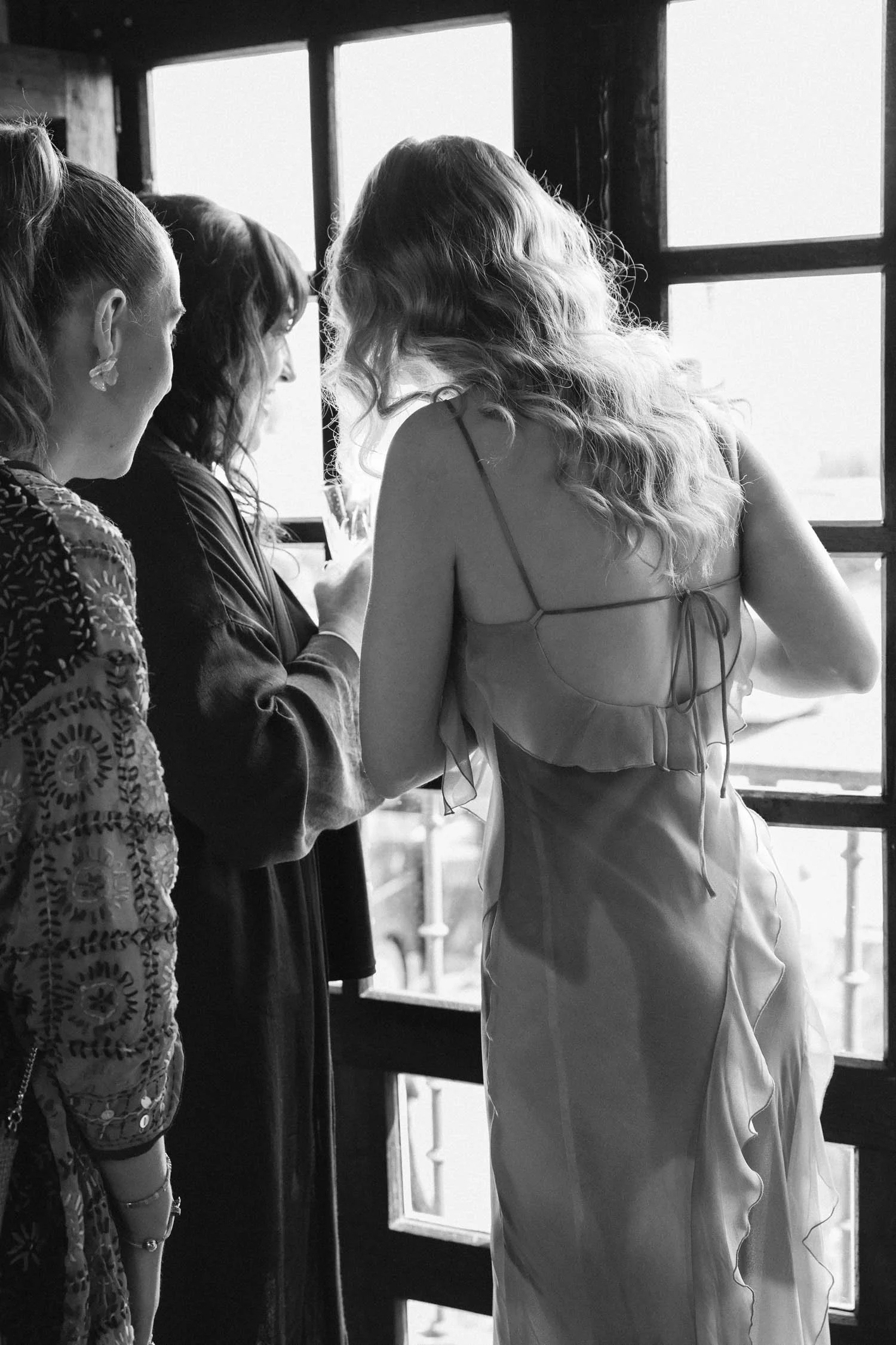 Two guests and bride are standing near a large window, with two of them closely looking at something in one woman's hand.  The scene is in black and white, indoors, with natural light coming through the window.