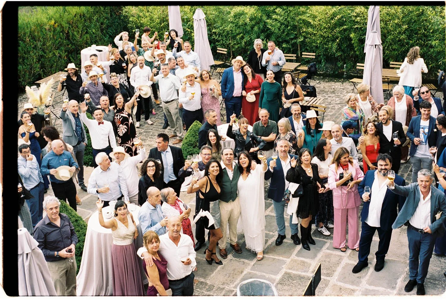A large group of people celebrating at an outdoor party with drinks, smiling, raising glasses, and mingling in a garden patio area.