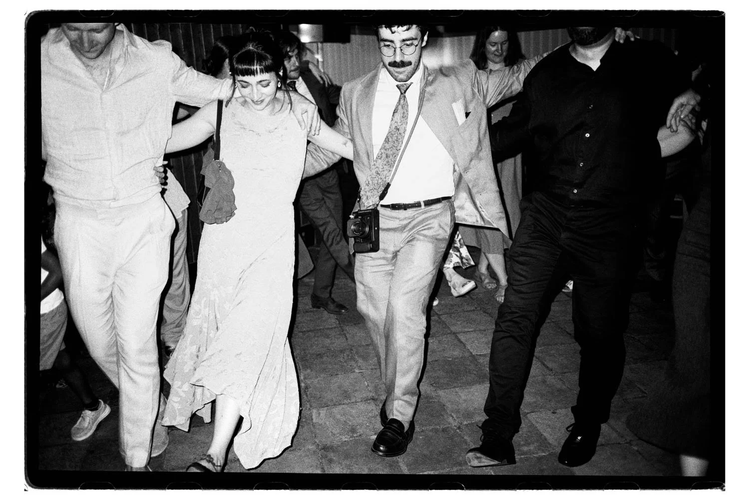 People dancing in a line at a social gathering, with some smiling and holding onto each other's shoulders.