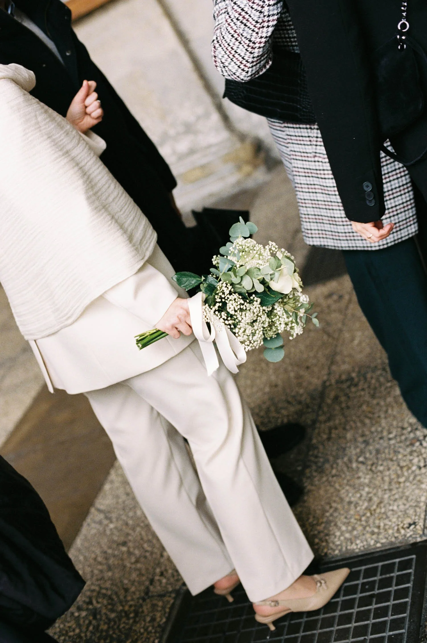 A woman in a white suit holding a bouquet of white flowers and greenery, standing next to a man wearing a plaid coat. They are at an indoor event, possibly a wedding.