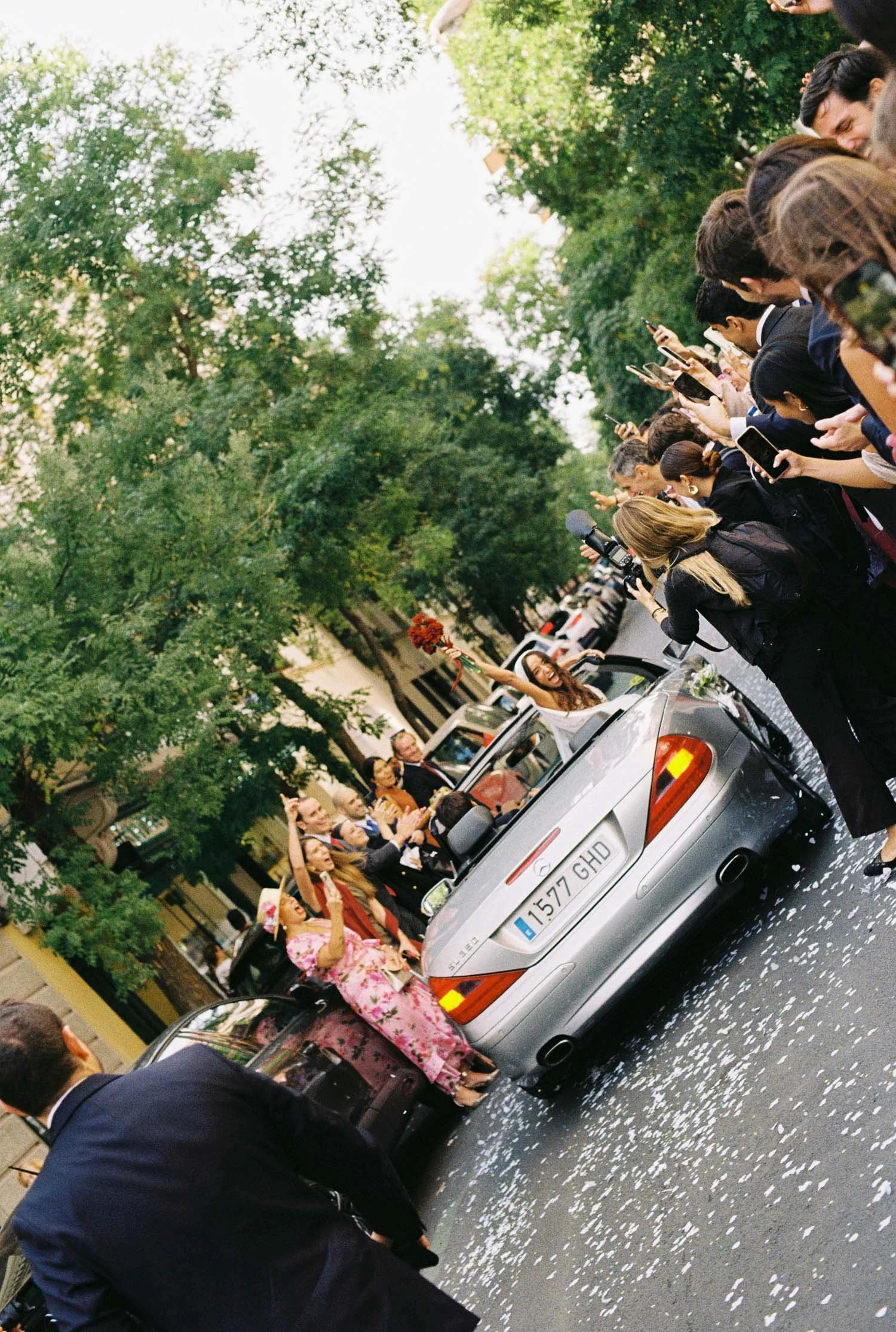 Group of women dressed in traditional Mexican dresses riding on top of cars during a parade or celebration. The women are waving, smiling, and taking photos, with a crowd of people and green trees on a city street in the background.