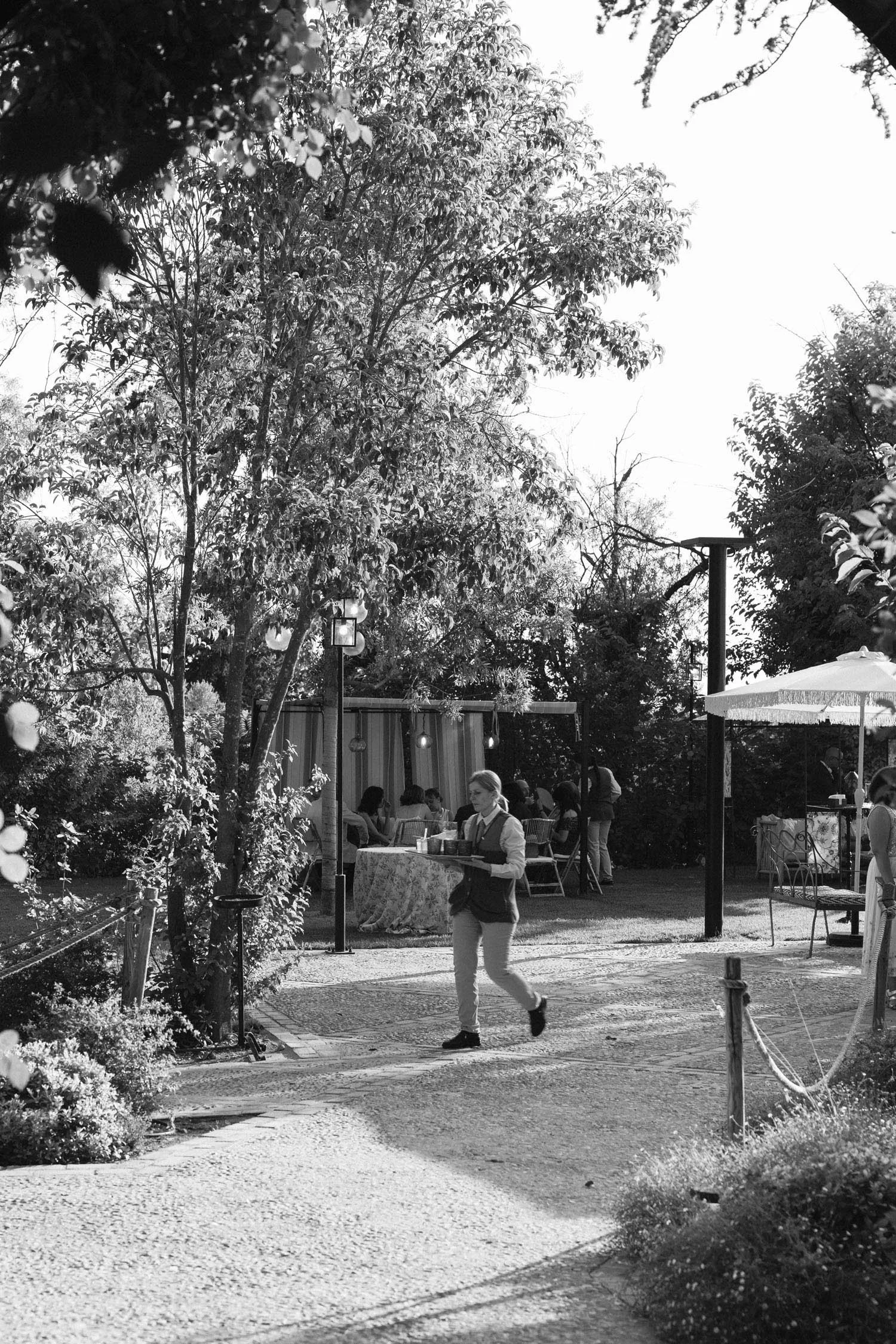 Black and white photo of an outdoor area with trees, a pathway, and a table with a tablecloth. A woman in a vest and pants is serving food, and several people are seated at the table under a canopy. Streetlights and outdoor furniture are also visible