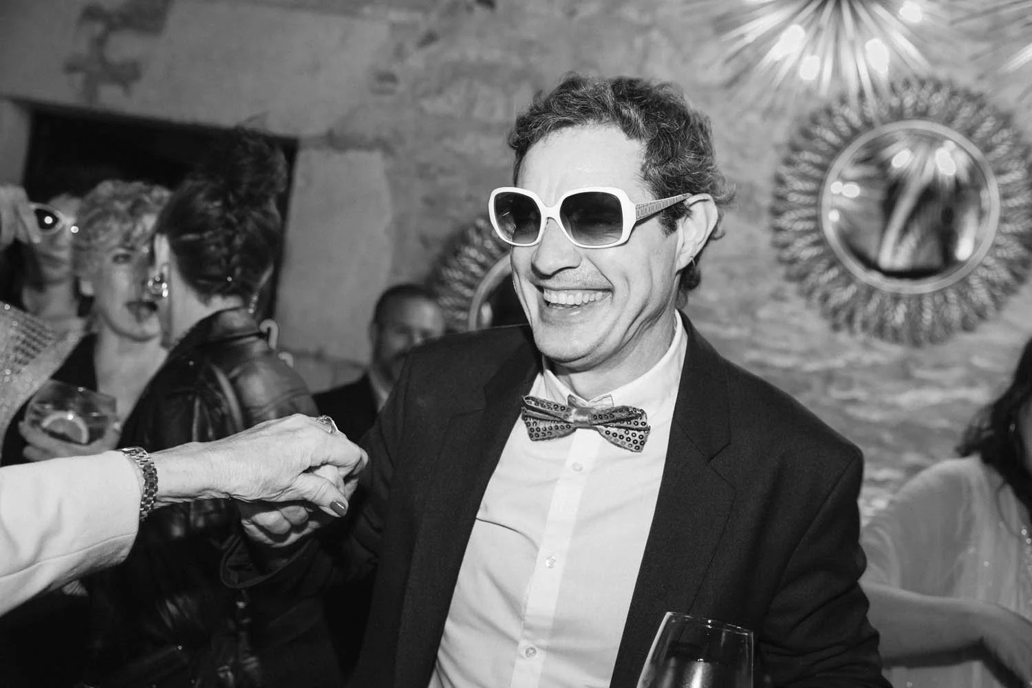 Black-and-white photo in a wedding party of a man wearing large sunglasses, a bow tie, and a suit, smiling while shaking hands.