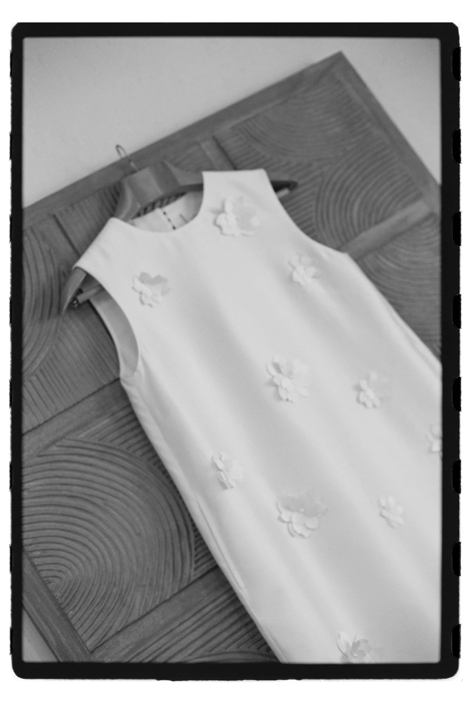 White dress with floral embellishments hanging on a wooden hanger on a patterned wooden surface.