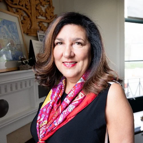 A woman with shoulder-length brown hair, wearing a black top and a colorful scarf, smiling in a well-lit room with artwork and decorative woodwork in the background.