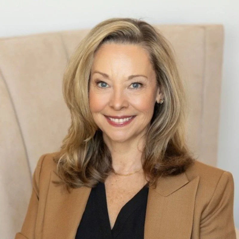 Portrait of a woman with shoulder-length blonde hair, blue eyes, and a bright smile, sitting on a beige headboard, wearing a tan blazer over a black top.