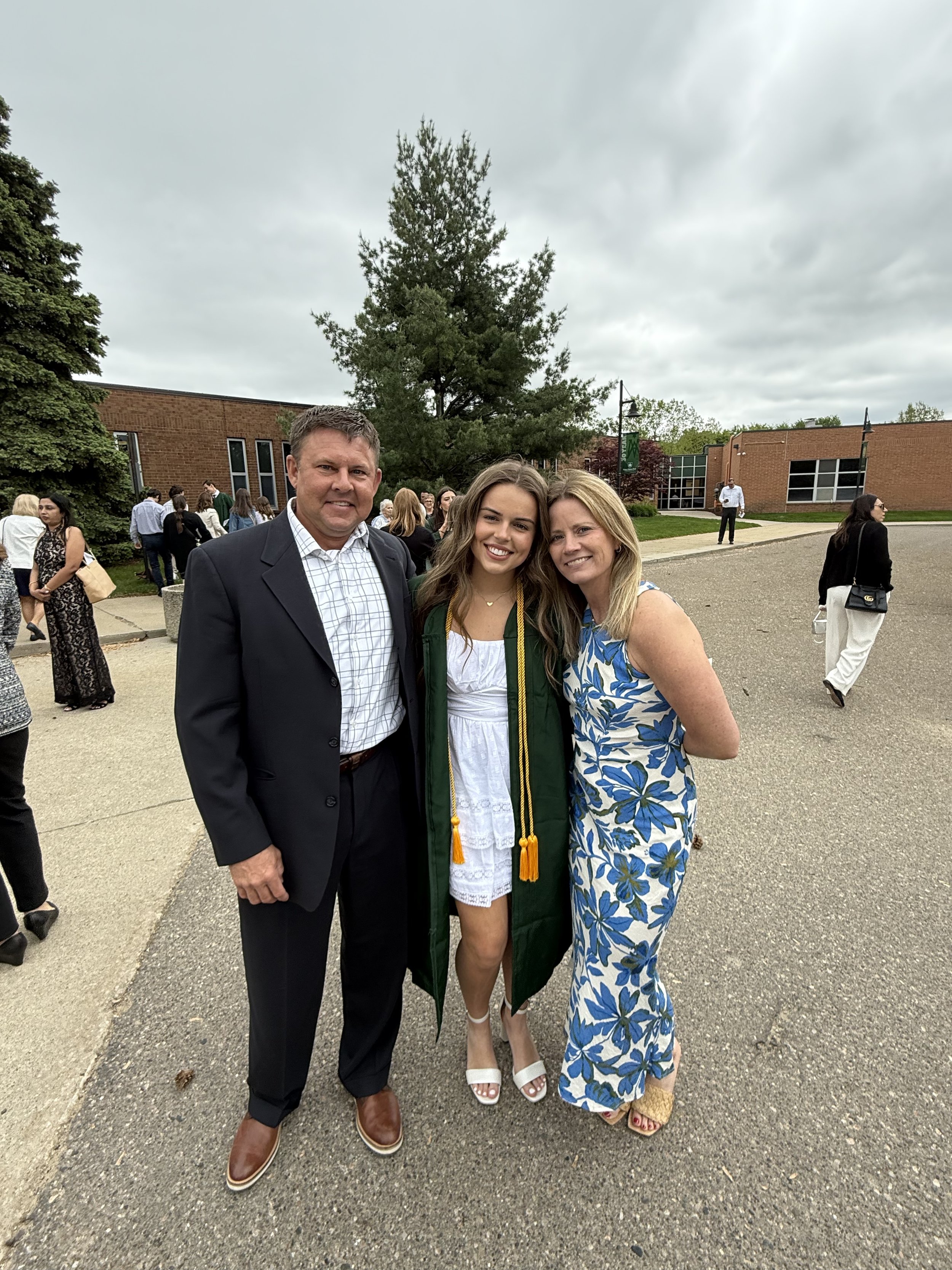 Three people posing outdoors at a graduation ceremony. The graduate, a young woman in the middle, wears a green gown and cap with a yellow honor cord, smiling. To her left, a man in a dark suit and dress shirt, and to her right, a woman in a floral dress, also smiling. Other attendees are visible in the background, with a brick building and trees under a cloudy sky.