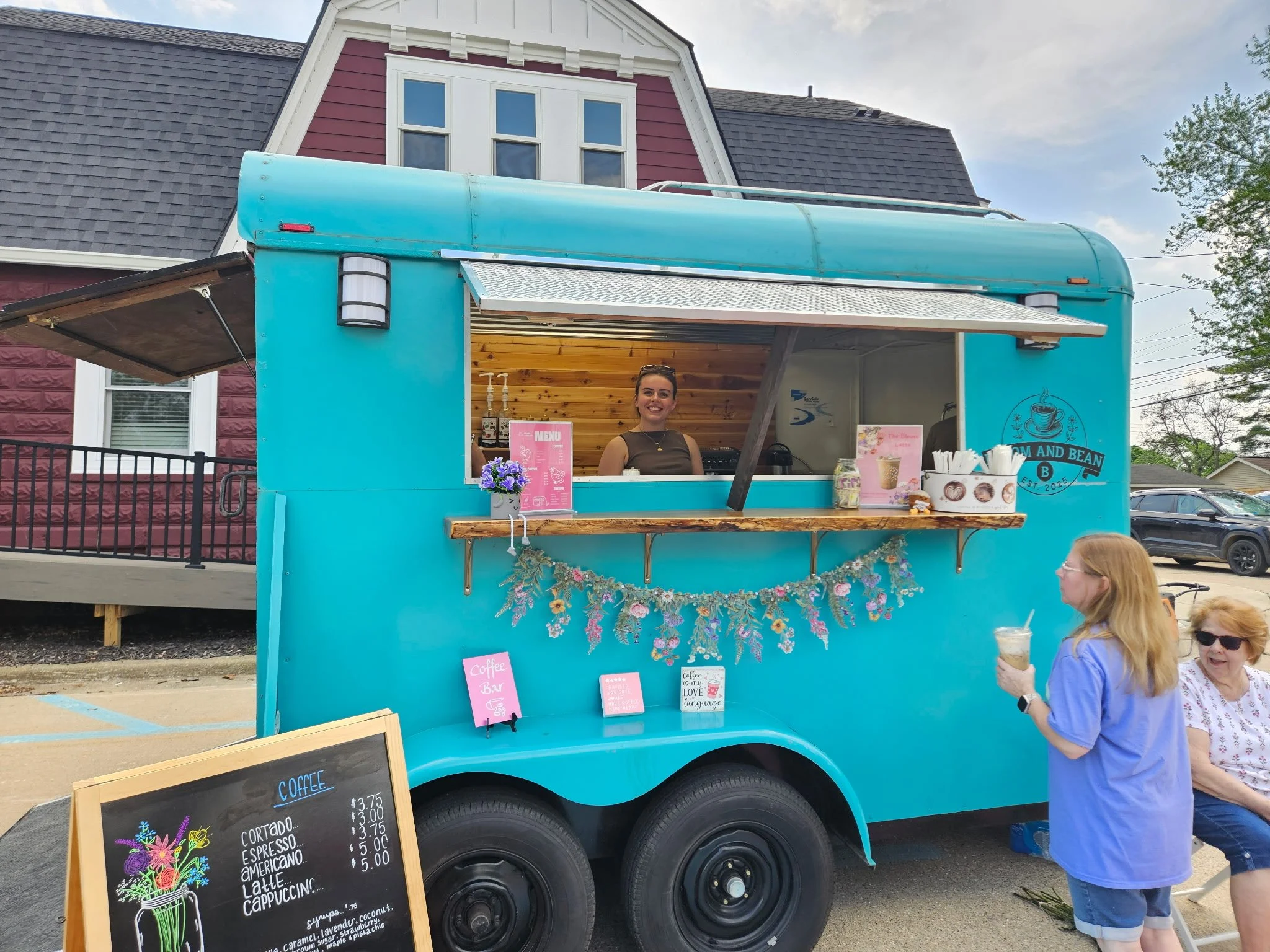A bright blue food truck serving coffee, with a woman smiling behind the counter. Outside, a woman in blue holds a coffee drink, and several people sit nearby. A blackboard menu displays coffee options and prices.