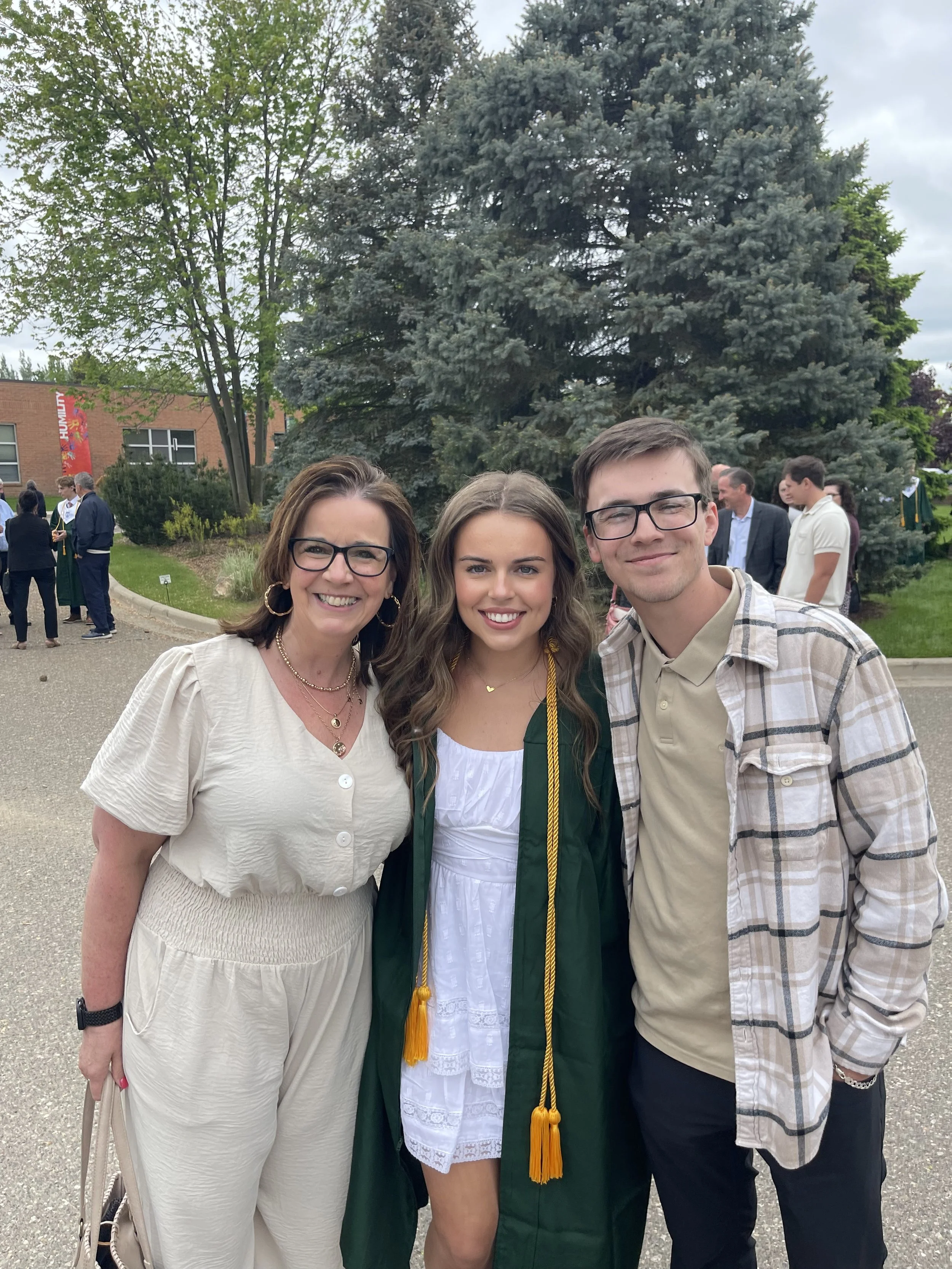 A woman in a white dress and glasses, a young woman in a green graduation gown, and a young man in a plaid shirt are smiling and posing together outdoors during a graduation celebration. There are other people and a large evergreen tree in the background.