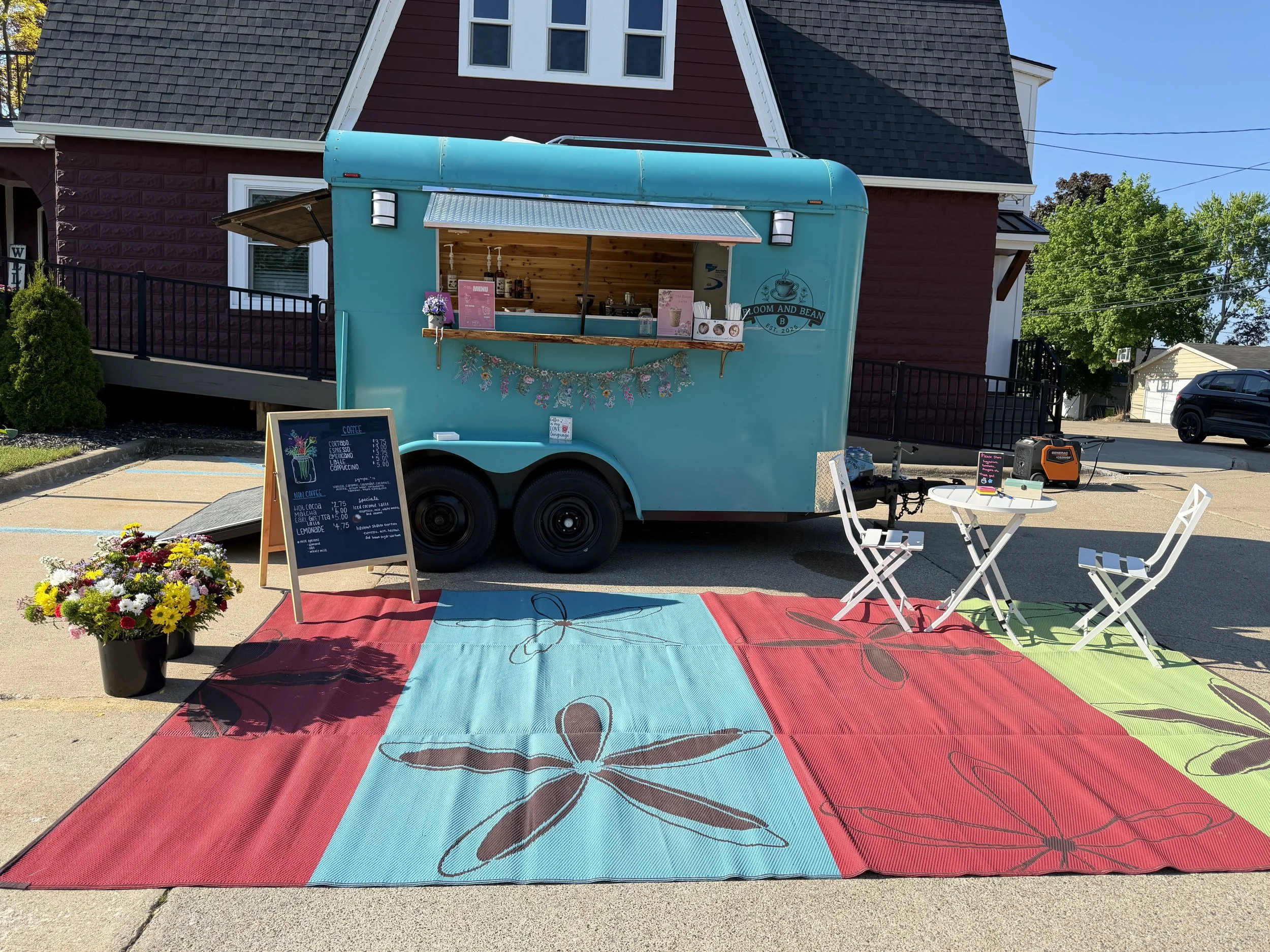 Blue food truck with a service window, menu, and floral decorations, parked in front of a red house with a black roof. Chairs and a table are set outside on a multicolored butterfly-patterned rug.