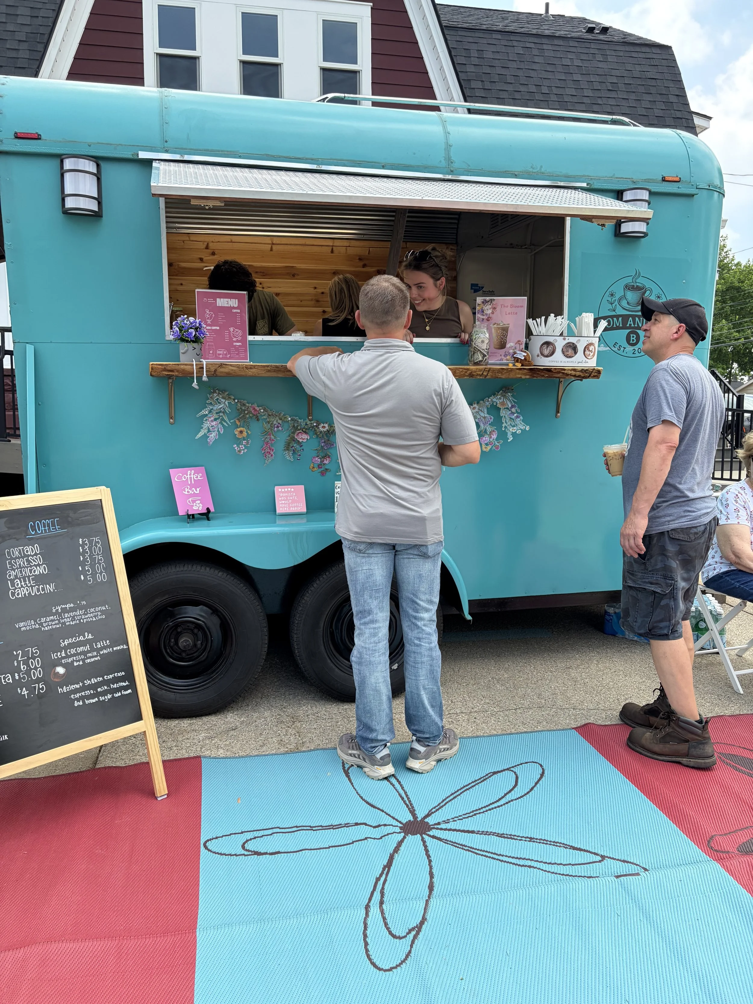 People ordering coffee at a teal food truck with a butterfly design on the side. The truck is parked on a street with a building in the background. There is a menu board and a colorful mat on the ground.