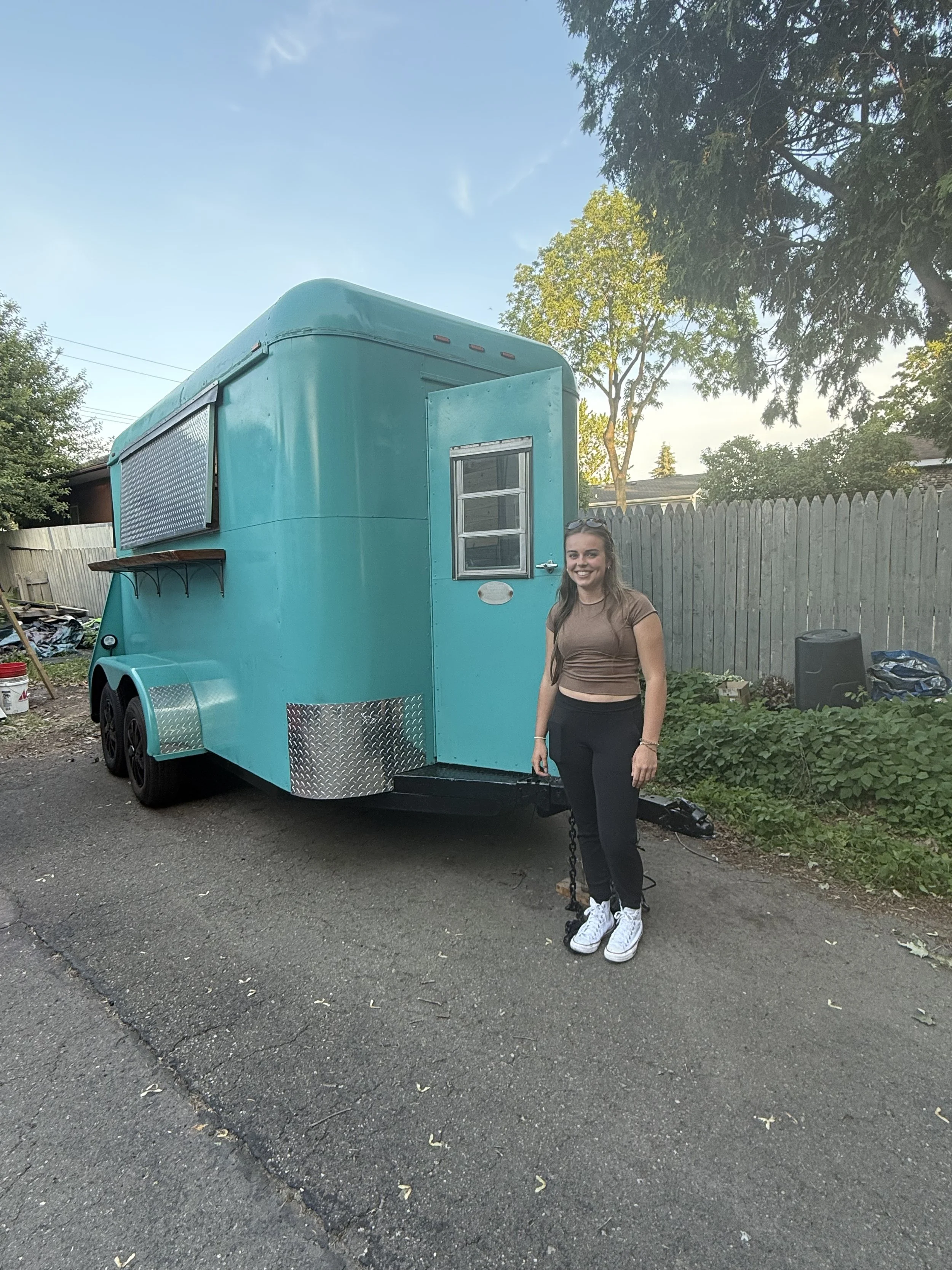 A smiling woman with long hair, wearing a brown crop top and black pants, stands outside next to a teal food trailer with a small window and a pull-out serving window, on a paved street with a wooden fence and trees in the background.