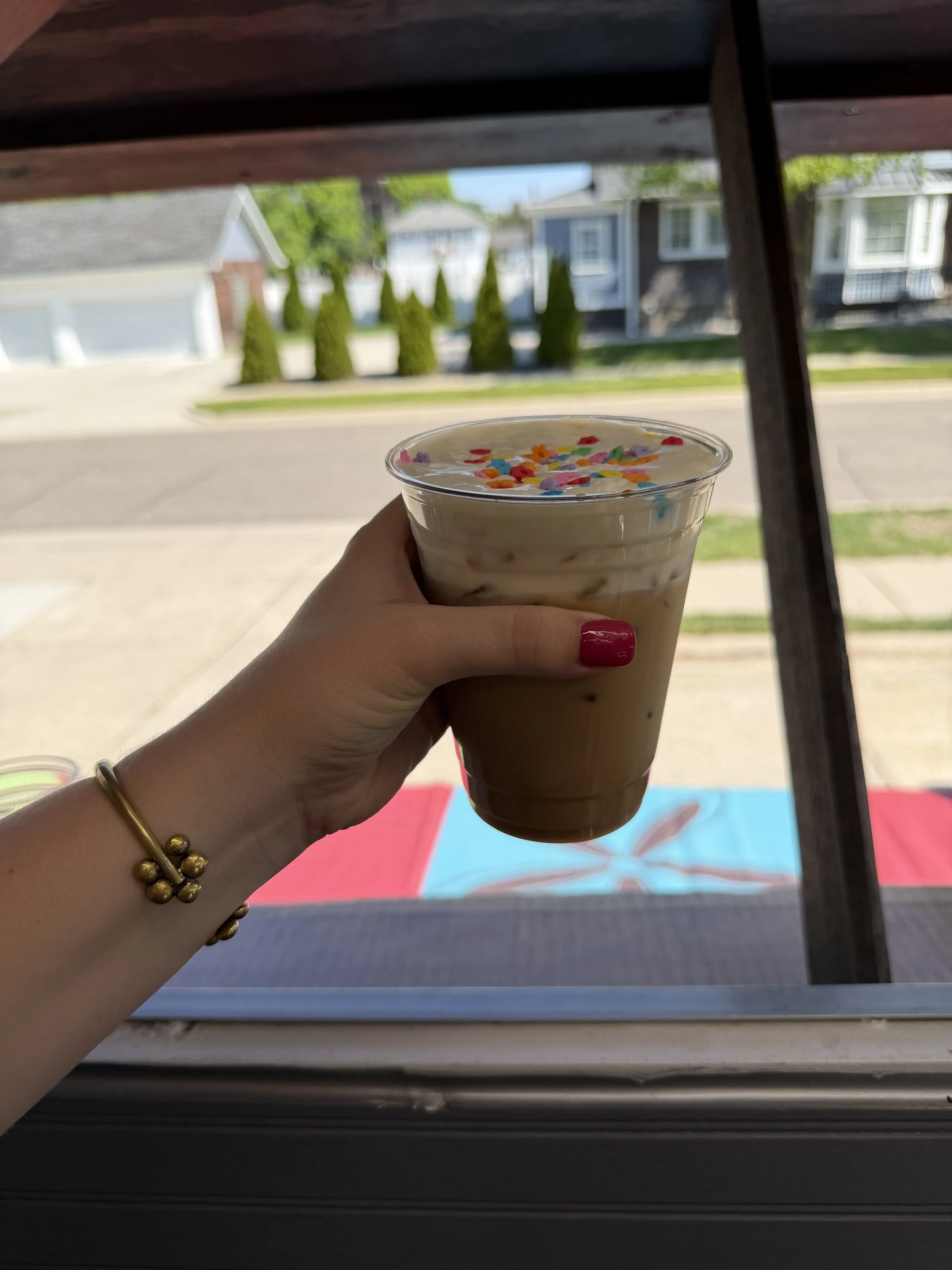 A hand with red painted nails holding a clear plastic cup filled with a light brown beverage topped with colorful heart and flower shaped sprinkles, seen through a window with blinds in a residential neighborhood during daytime.