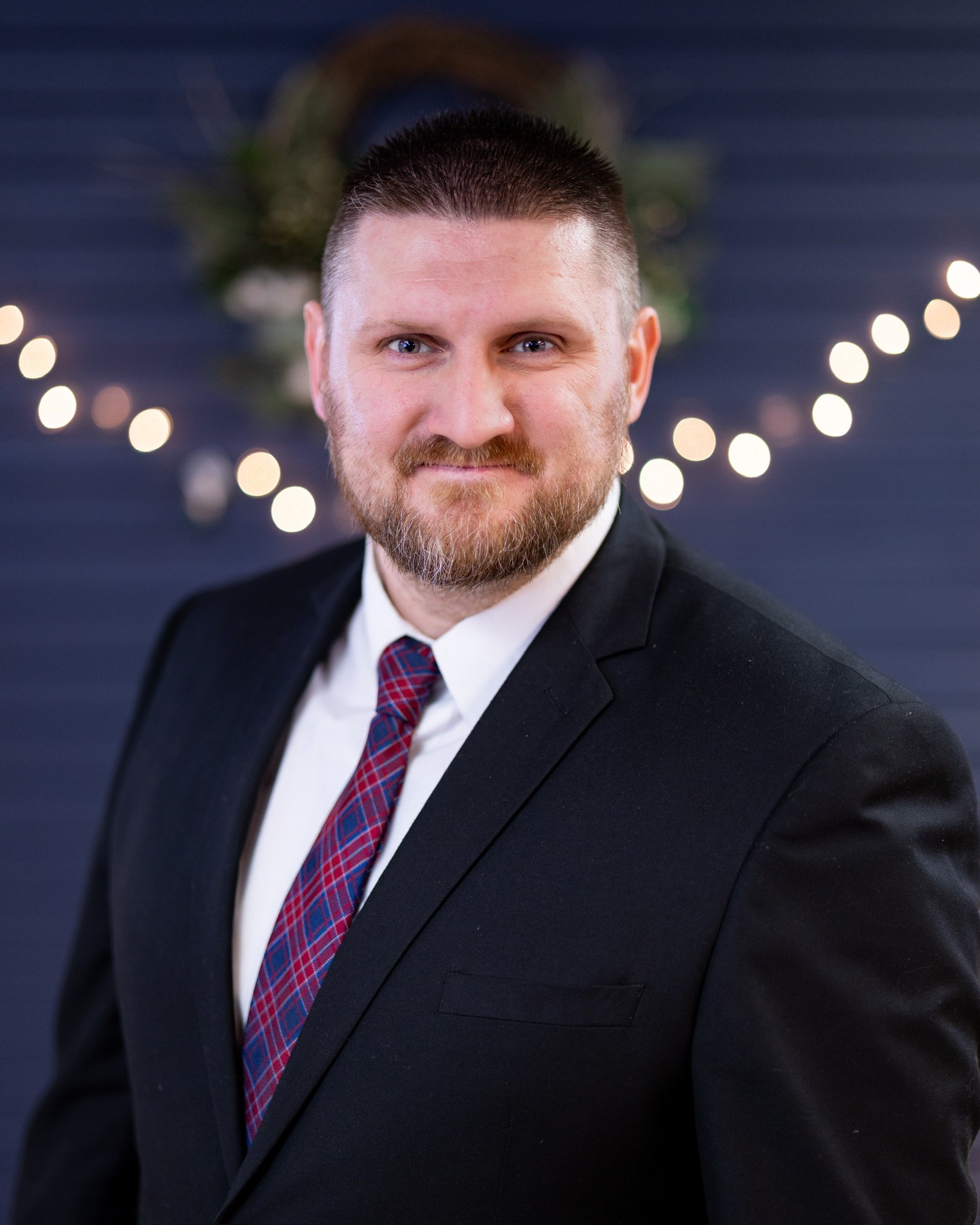 A man with a beard and short hair wearing a black tuxedo, white shirt, and a red & blue checkered tie, smiling at the camera with a blurred background of Christmas lights and decorations.