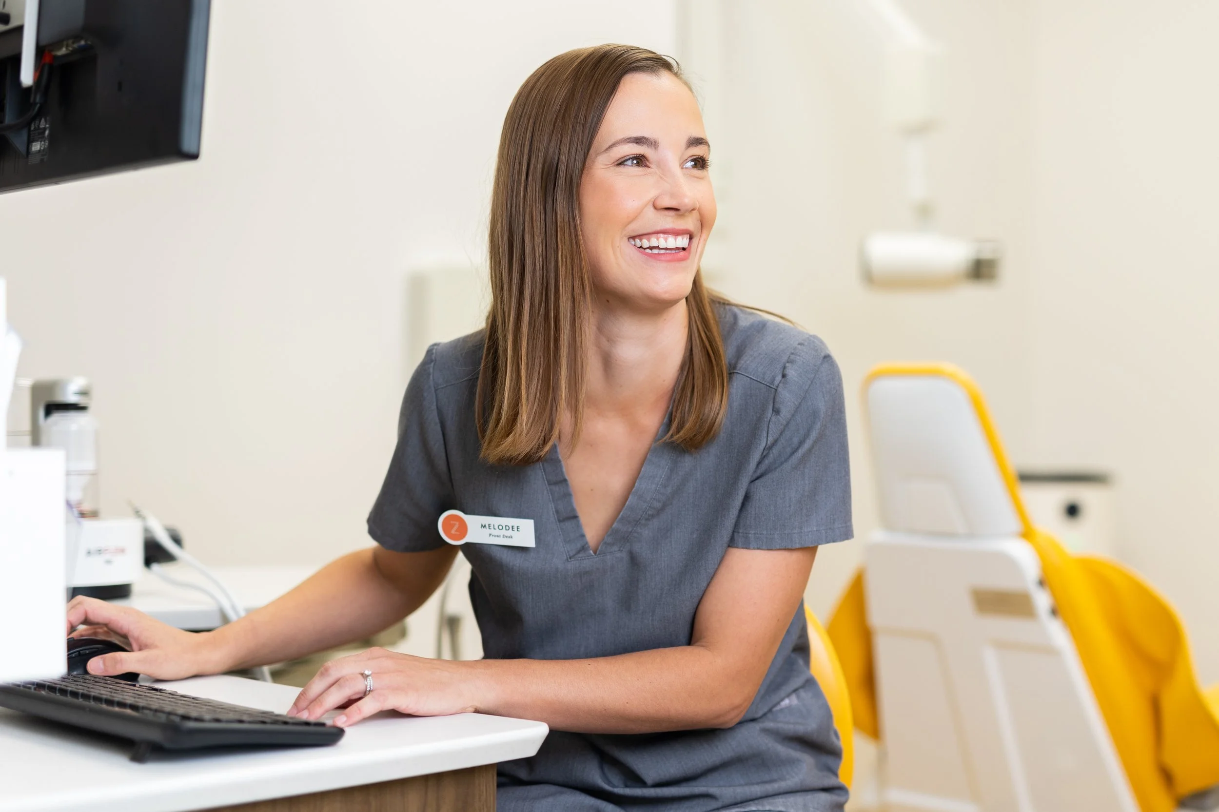A smiling woman with long brown hair, wearing a gray shirt with a name tag that reads 'Melodee,' working on a computer in a medical or office setting.