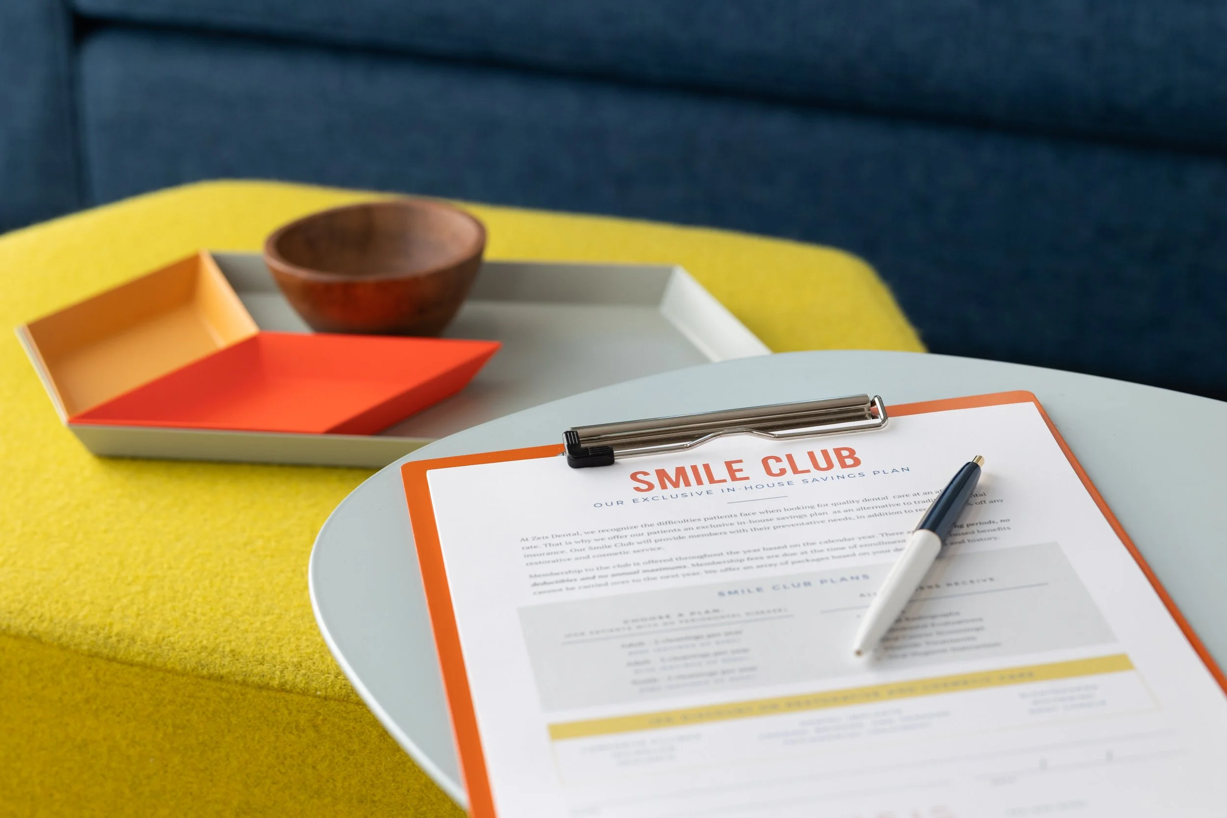 A table with a Smile Club plan document, a silver and white pen, a tray with a wooden bowl and colored paper trays, on a yellow tablecloth and a blue sofa in the background.