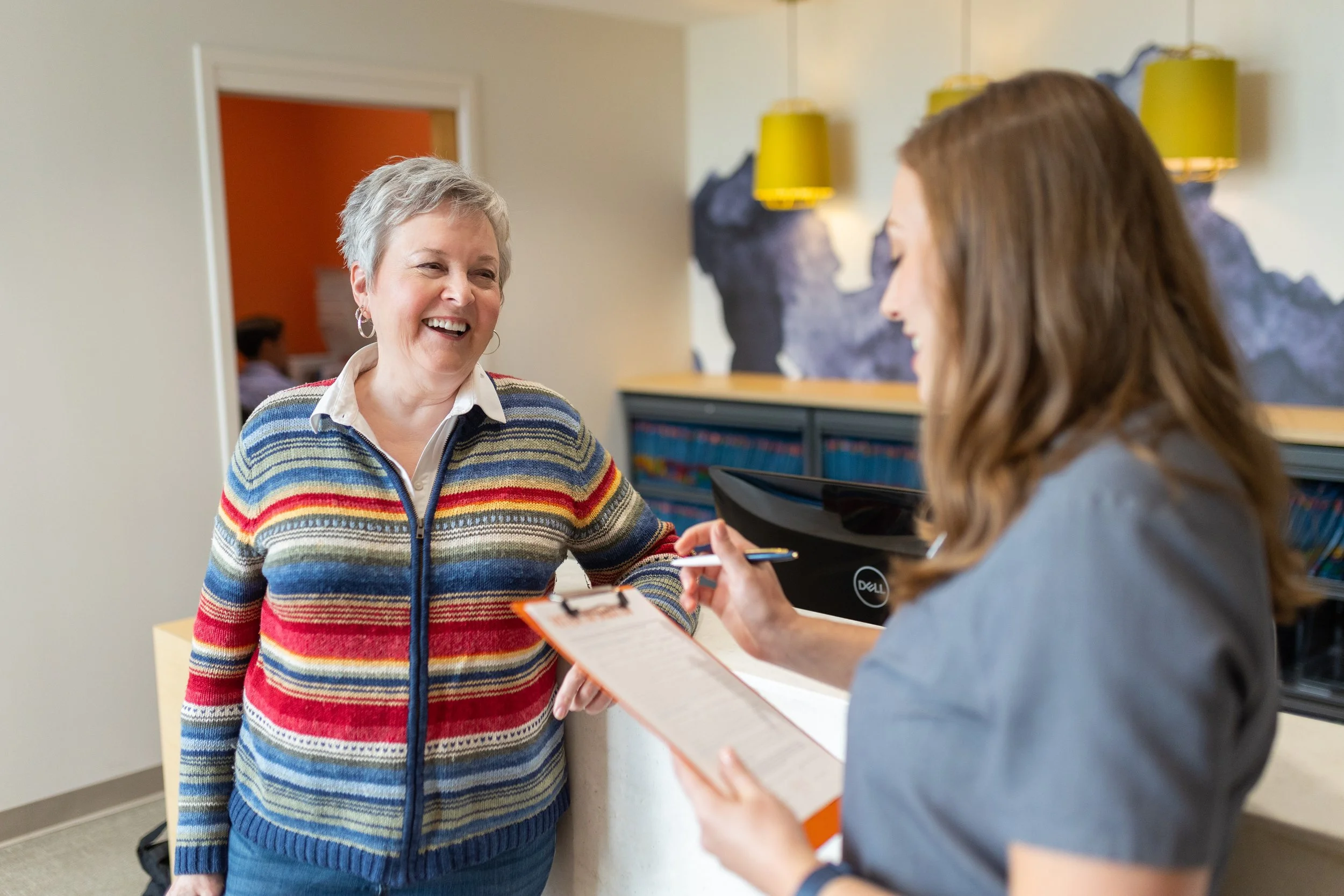 An older woman in a colorful striped sweater smiling during a conversation with a younger woman holding a clipboard in an office setting, with artwork and books visible in the background.