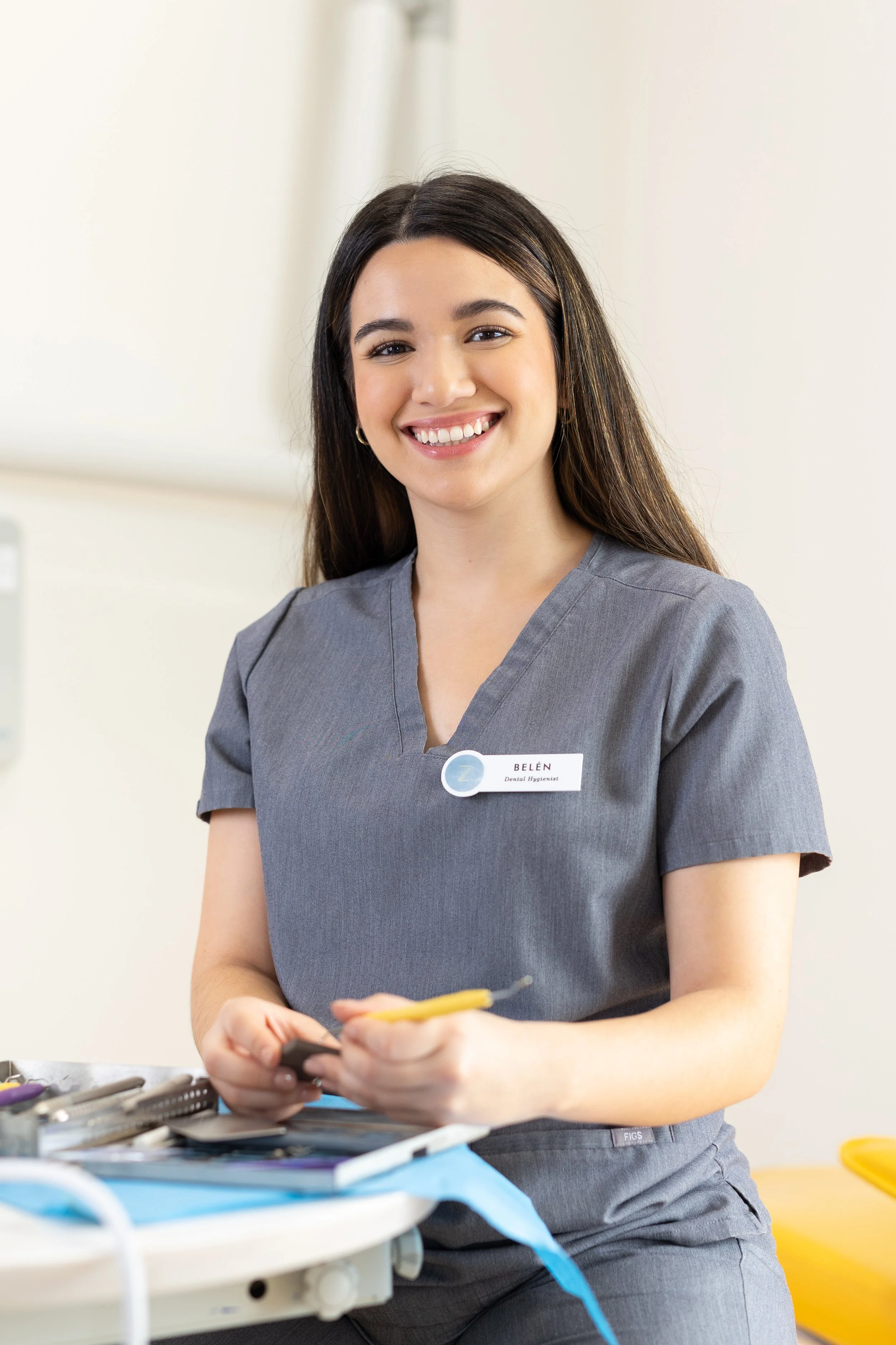 A young woman with long dark hair smiling in a gray medical uniform, standing in a clinical setting with dental tools on the table in front of her.