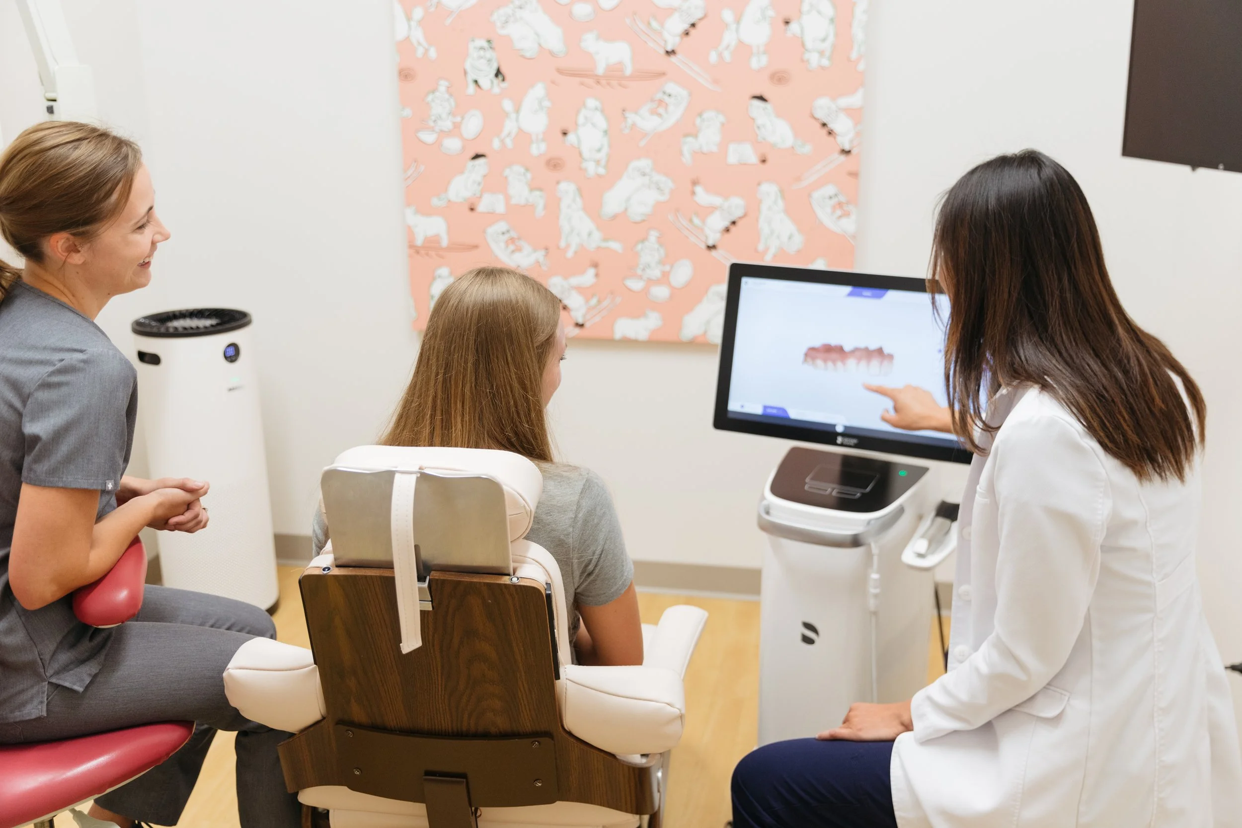 A girl sitting in a dental chair with a dental assistant and a woman, possibly a mother, sitting beside her. The dental assistant is pointing at a computer screen displaying dental x-ray images. The woman is smiling, and the background features a wall with a pink pattern of white dogs.