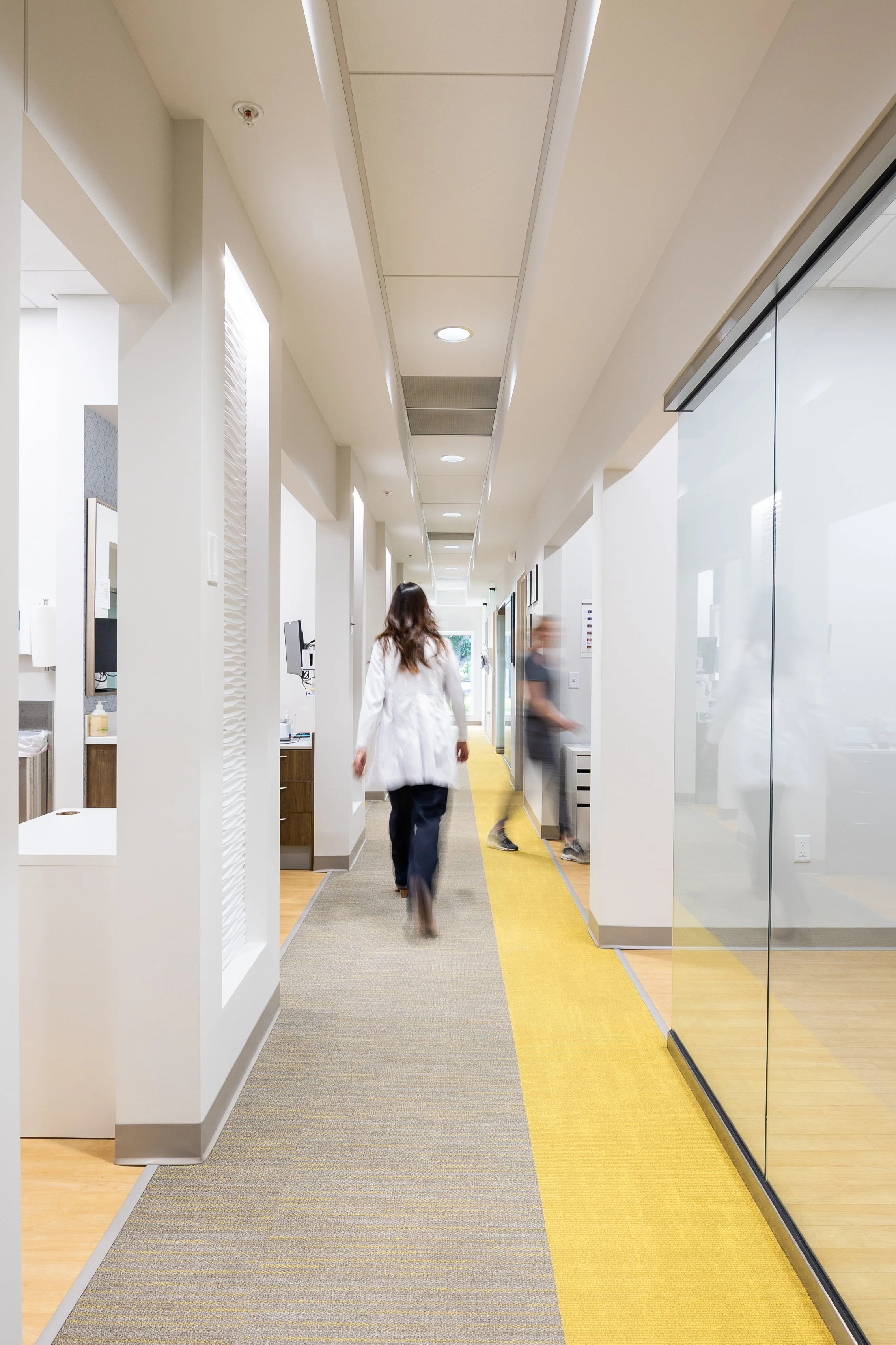 Hospital corridor with medical staff walking and various rooms visible.