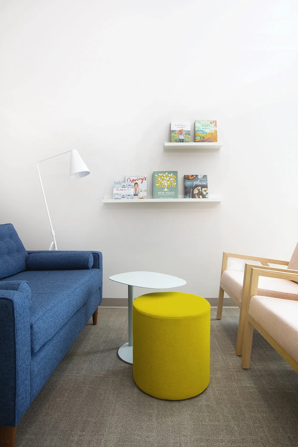 Cozy waiting room with blue sofa, pink armchair, white side table, yellow ottoman, white floor lamp, and wall-mounted white shelves with books.