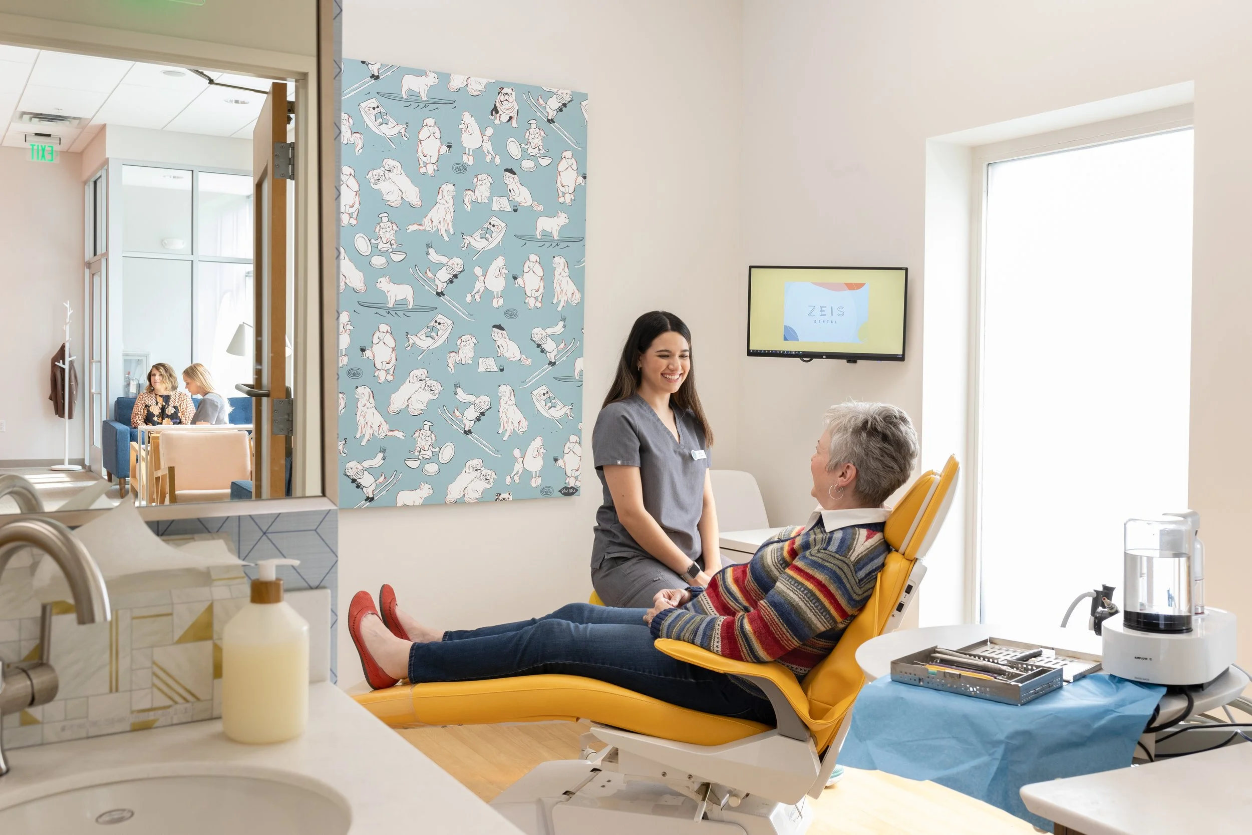 A woman receiving a dental consultation from a dental hygienist in a dental clinic. The patient is sitting in a dental chair, smiling, wearing a colorful striped sweater and red shoes. The hygienist is standing beside her, smiling, in gray scrubs. Th
