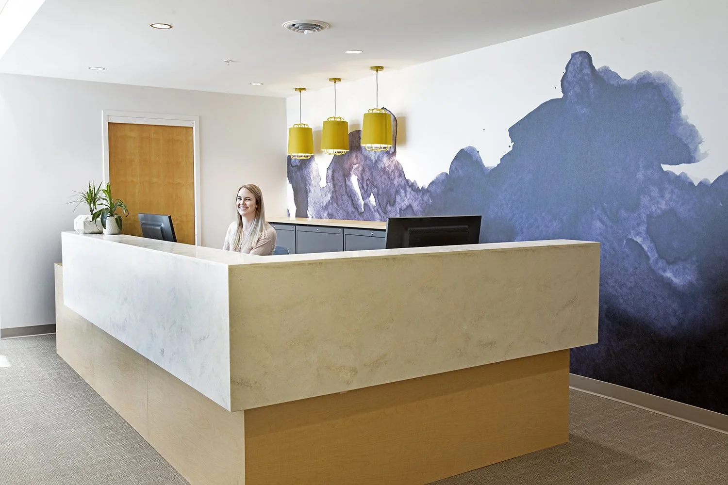Reception desk in an office with a smiling woman sitting behind it, a painting of mountains on the wall, yellow pendant lights hanging from the ceiling, and potted plants nearby.