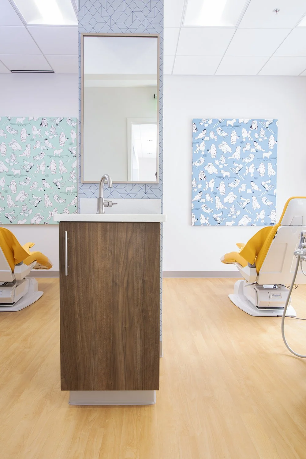 Dental exam room with yellow dental chairs, artwork with animals on the walls, a mirror, a wooden cabinet with a sink, and a partition in the center.