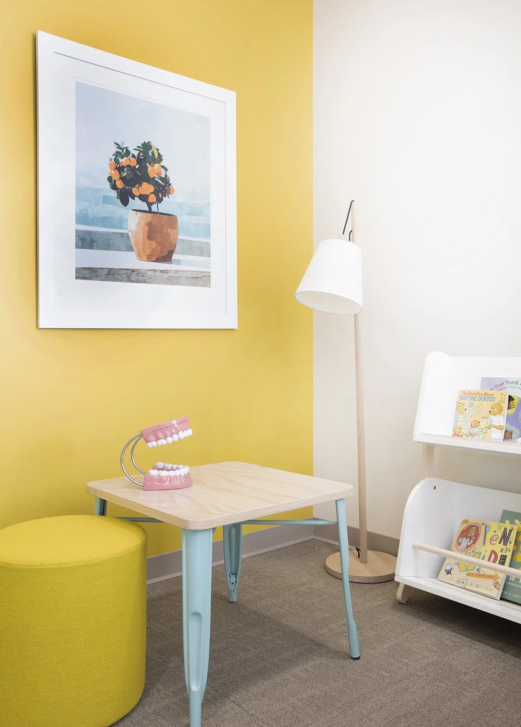 A children's dentist office with a small wooden table, a pink dental model, a yellow stool, a white bookshelf with children's books, a white floor lamp, and framed artwork of a potted plant on a yellow wall.