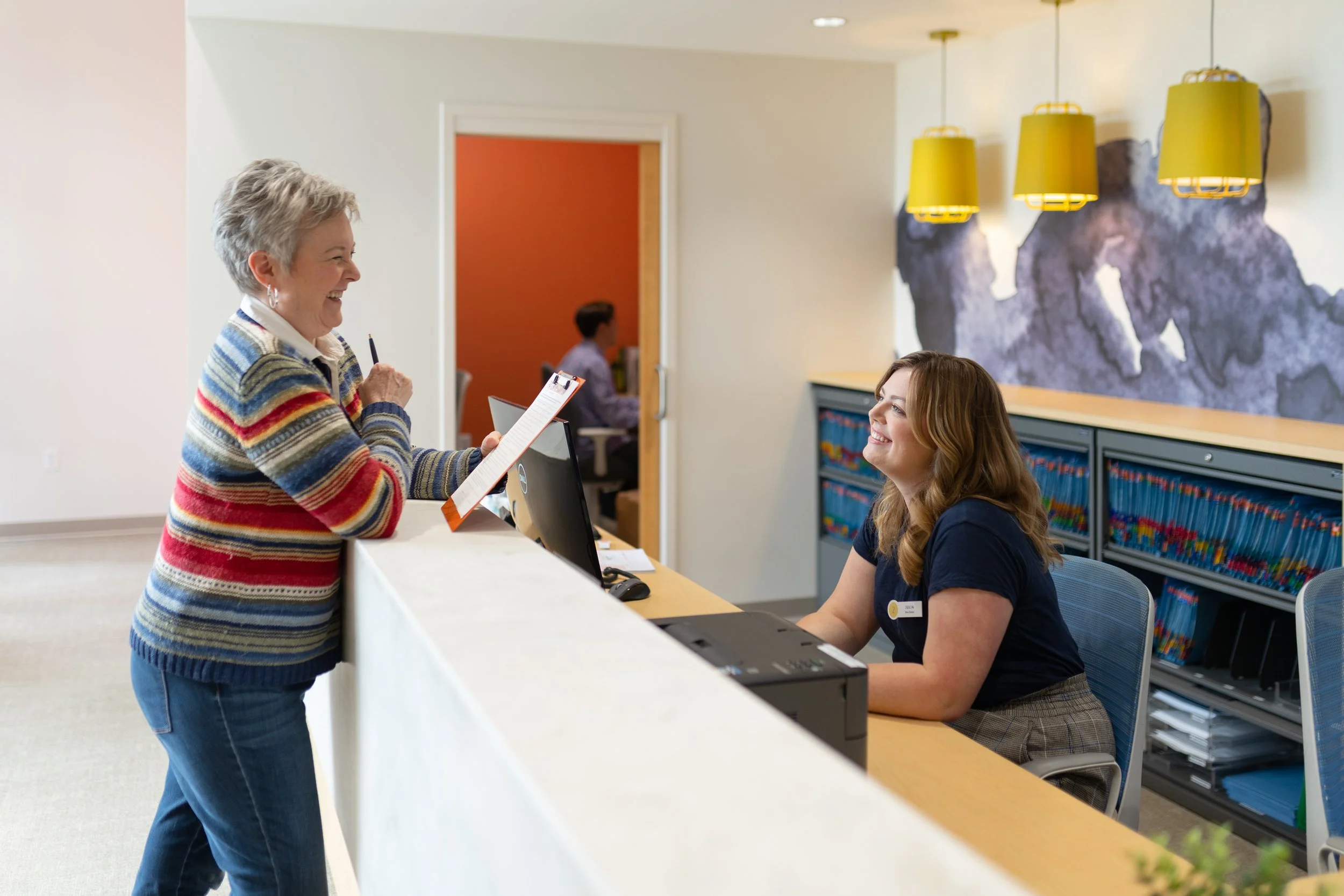 A woman at a reception desk talking to a receptionist. The woman is smiling and holding a clipboard, while the receptionist is sitting behind the desk smiling back. The reception area has yellow pendant lights and a modern wall art.
