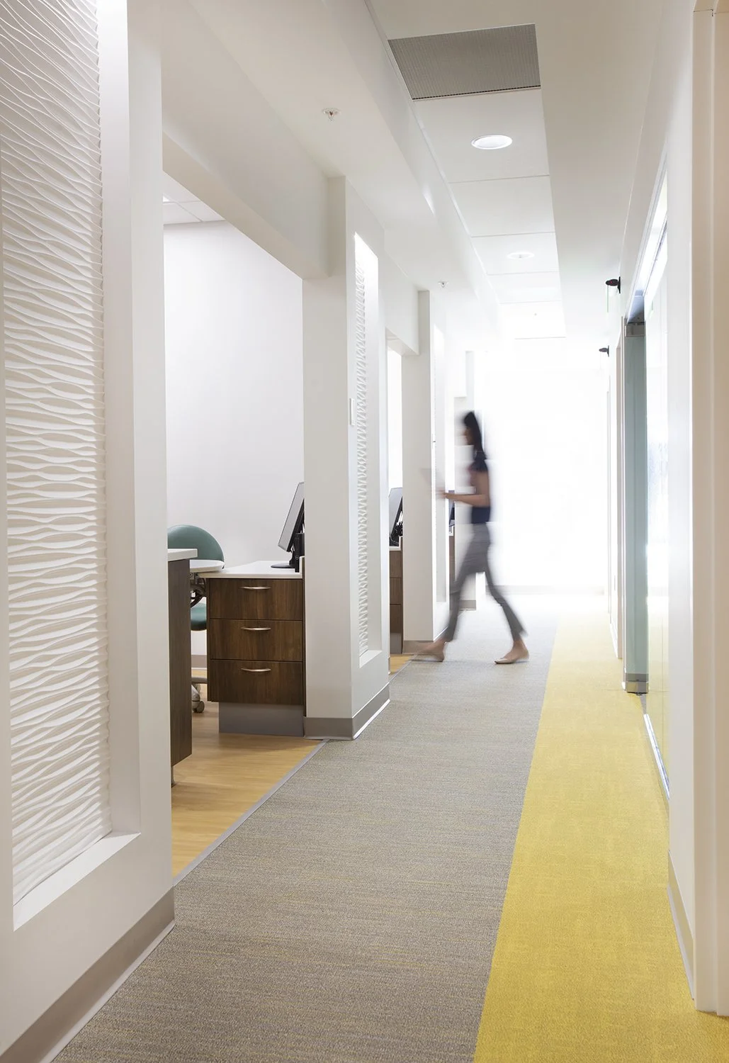 Office corridor with small office cubicles, one woman walking past, computer desks, and chairs, in a modern, well-lit office space.