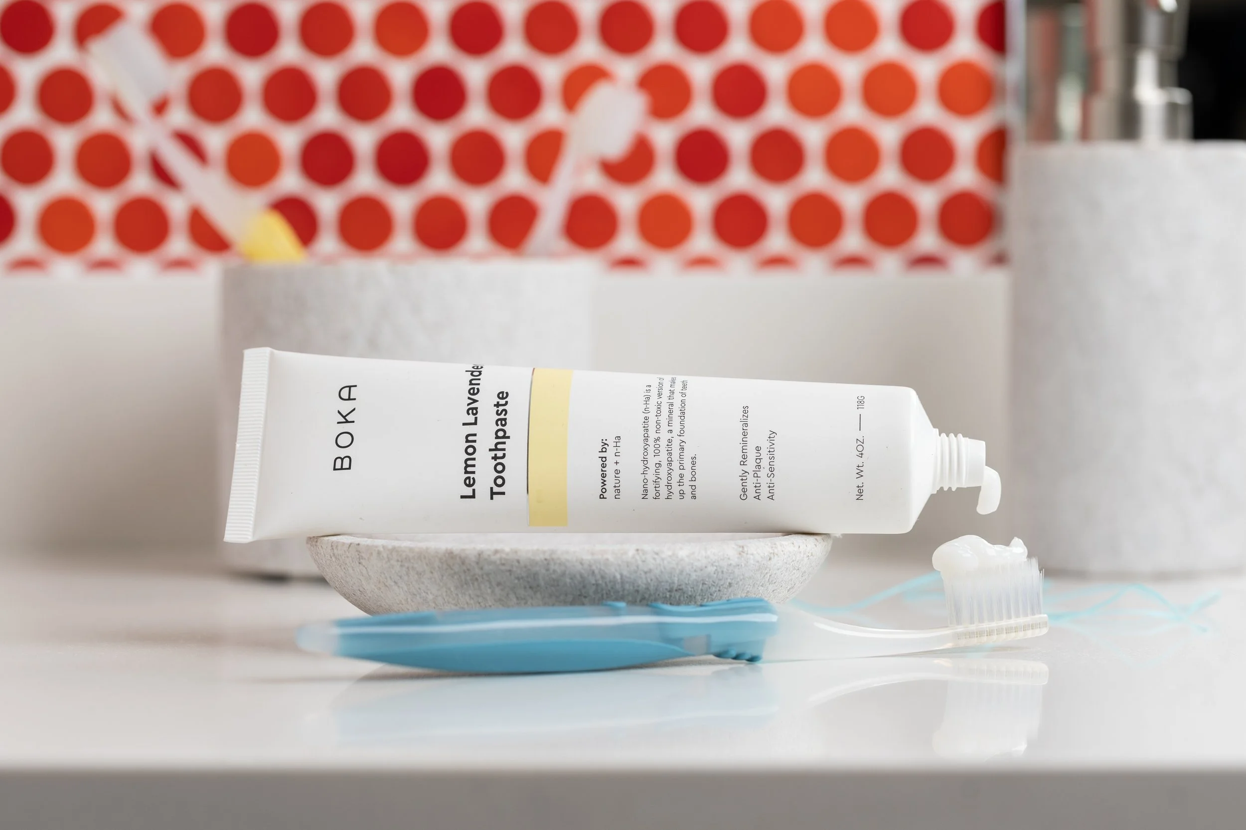 A tube of lemon lavender toothpaste resting on a small white dish with a toothbrush nearby; bathroom items in the background.