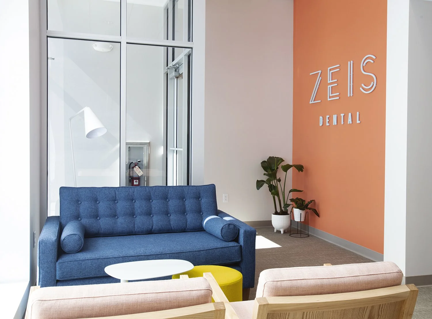 Waiting area inside Zion's Dental office with a blue sofa, pink chairs, a white coffee table, a yellow ottoman, a potted plant, and a pink accent wall with the dental office's name displayed.