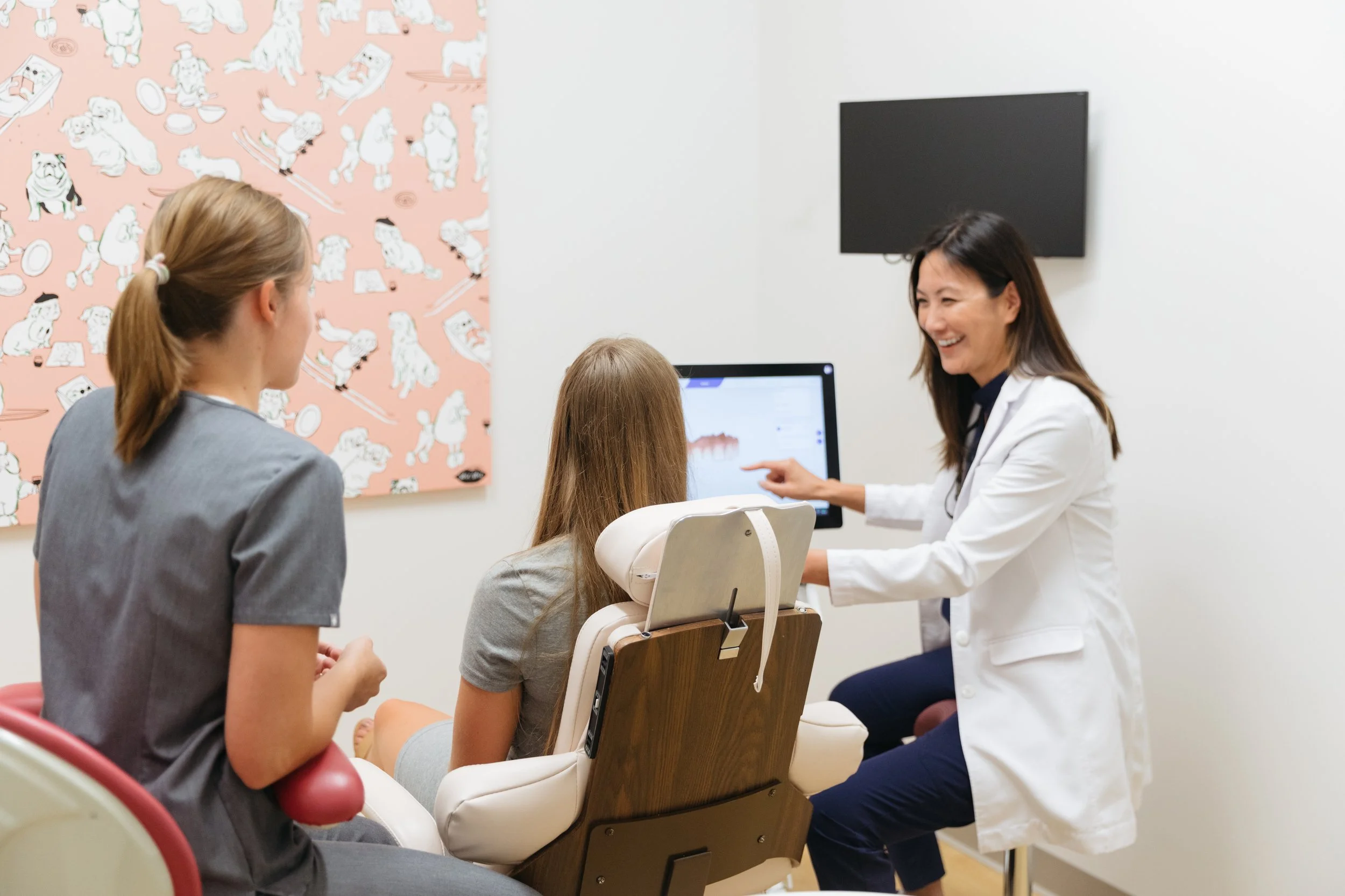 A female doctor in a white coat talking to a girl sitting in a dental chair, with two other women nearby, in a clinical setting with animal-themed wall art.