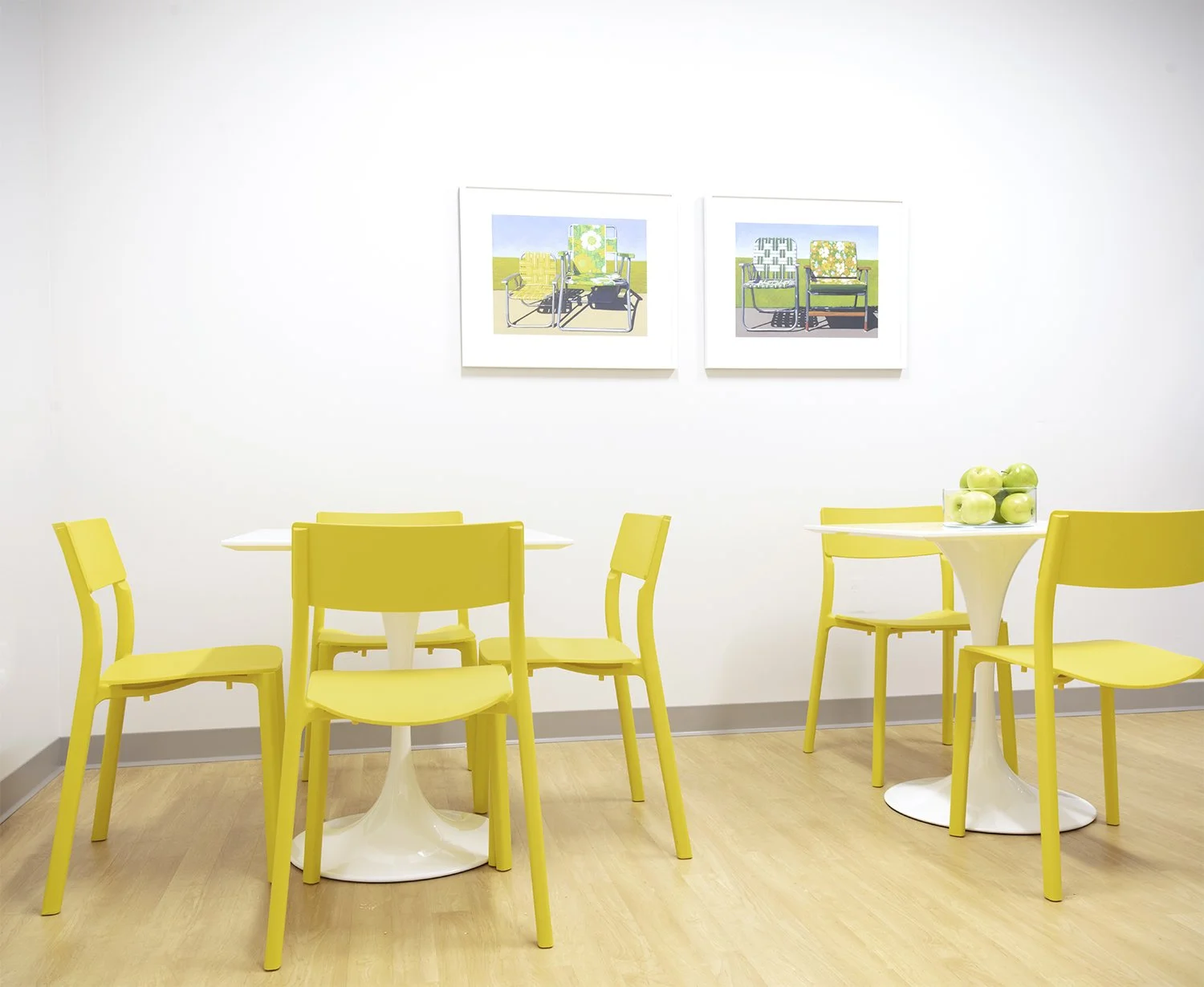 A brightly lit room with yellow chairs and white tables, decorated with a bowl of green apples, and two pieces of framed artwork on the wall featuring colorful outdoor furniture.
