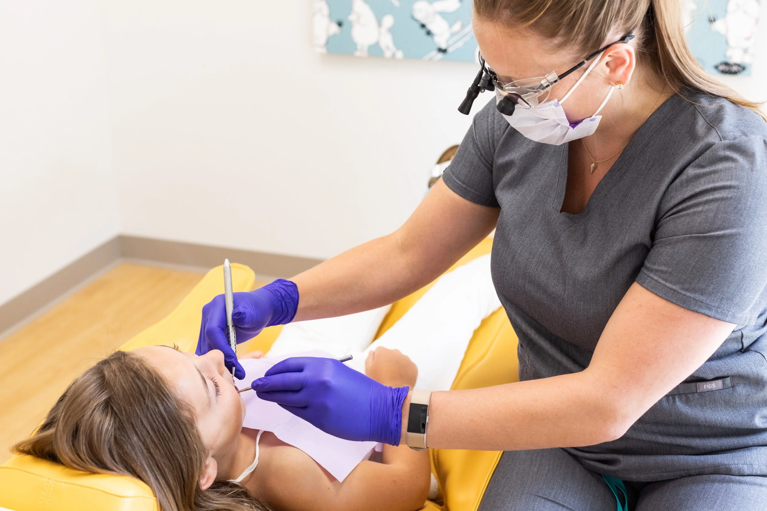 Dentist wearing dental loupes and mask treating a girl patient lying on dental chair, using dental instruments while the patient is seated with mouth open.