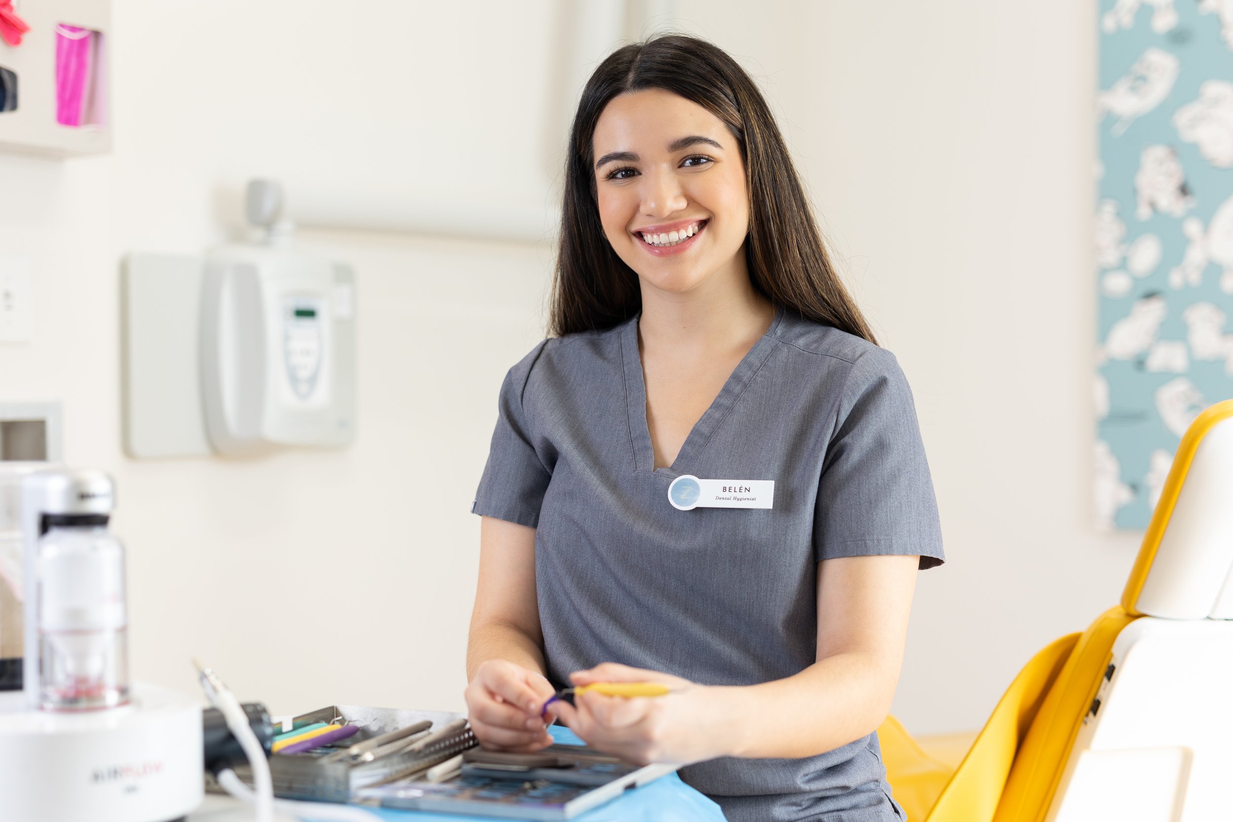 A smiling female dental professional in gray scrubs sitting at a dental clinic, holding dental tools, with dental equipment and supplies on the counter nearby.