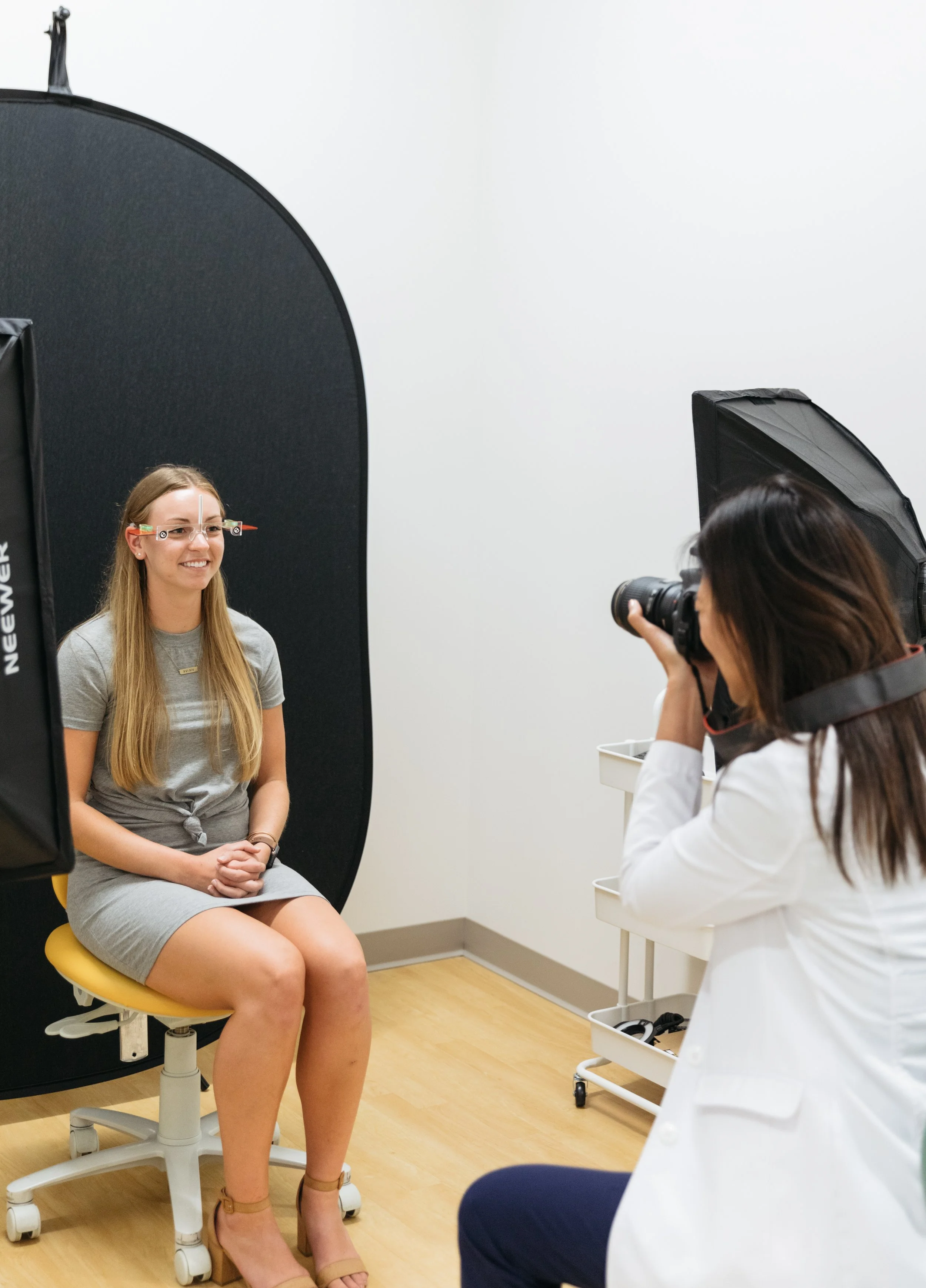 A woman with long blonde hair and glasses is sitting on a yellow stool, smiling, during a photoshoot or interview. A female photographer with dark hair and a white coat is taking her picture with a camera. The setting appears to be a photography studio with black backdrop and lighting equipment.