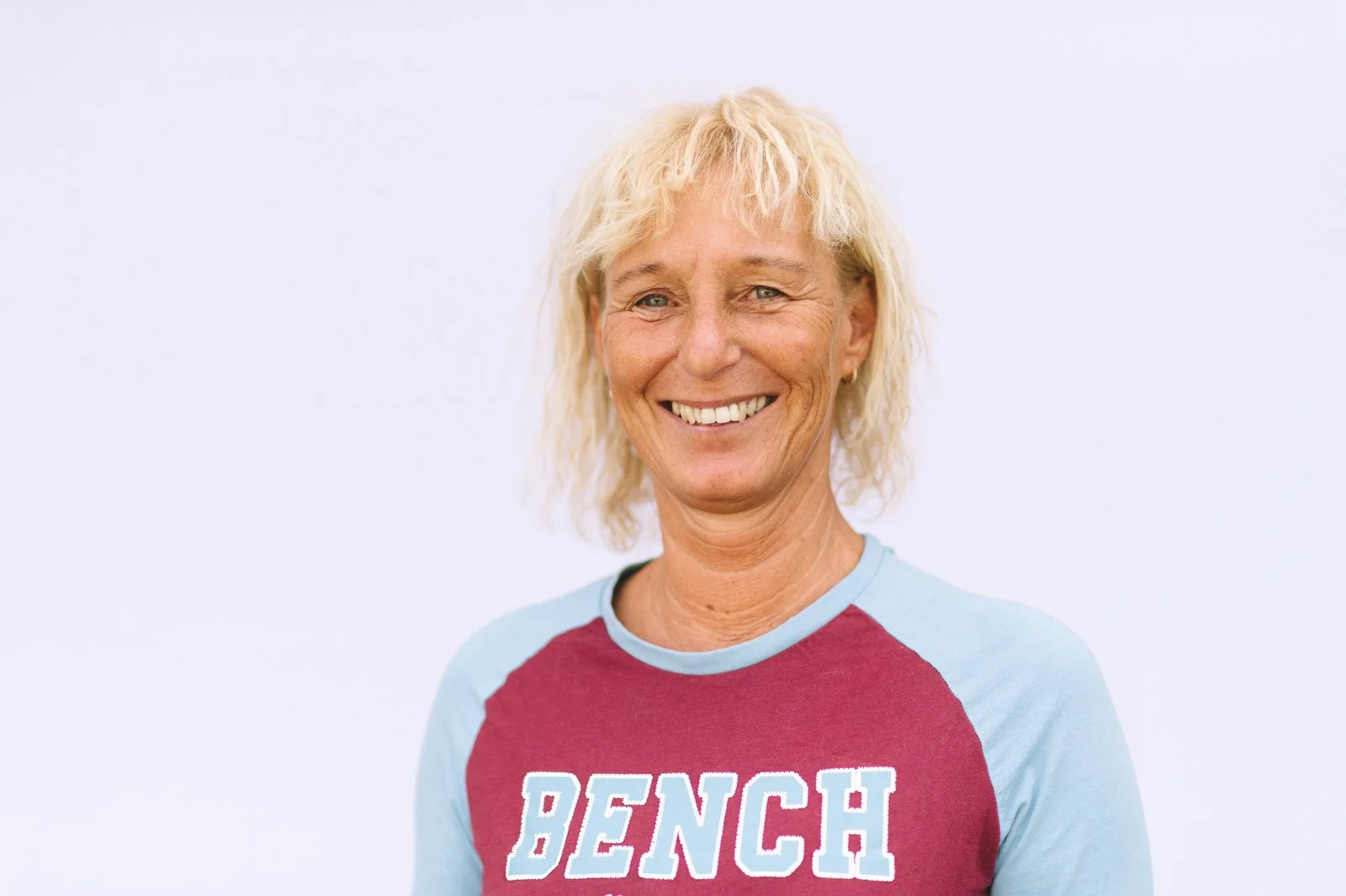 Eine lachende ältere Frau mit blonden, lockigen Haaren trägt ein rotes T-Shirt mit blauen Ärmeln, auf dem das Wort 'BENCH' steht, vor einem weißen Hintergrund.