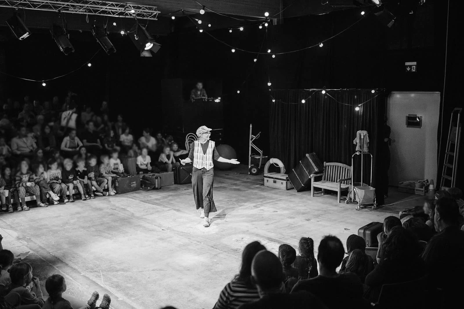 Ein Straßenkünstler auf einer Bühne, die von einem Publikum beobachtet wird, im Hintergrund hängen Lichter. Der Künstler trägt eine Mütze und eine Weste, während er eine Show präsentiert.