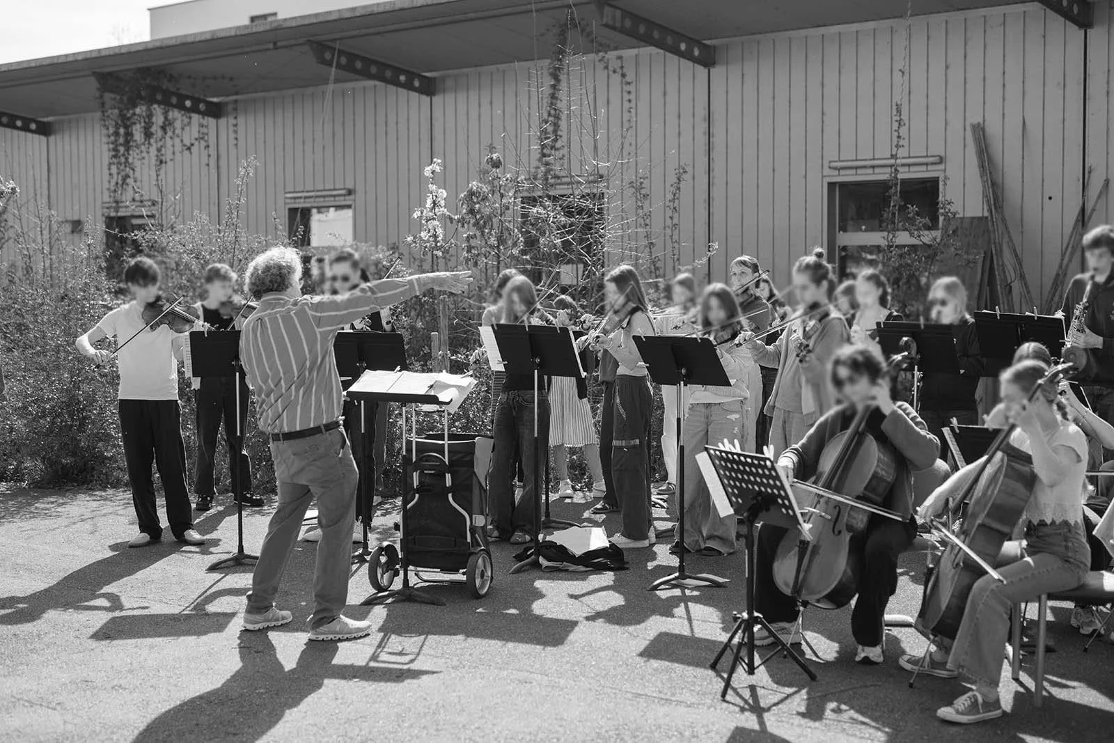 Ein Orchester mit mehreren Kindern und einem Dirigenten, das im Freien vor einem Gebäude mit Holzfassade spielt.