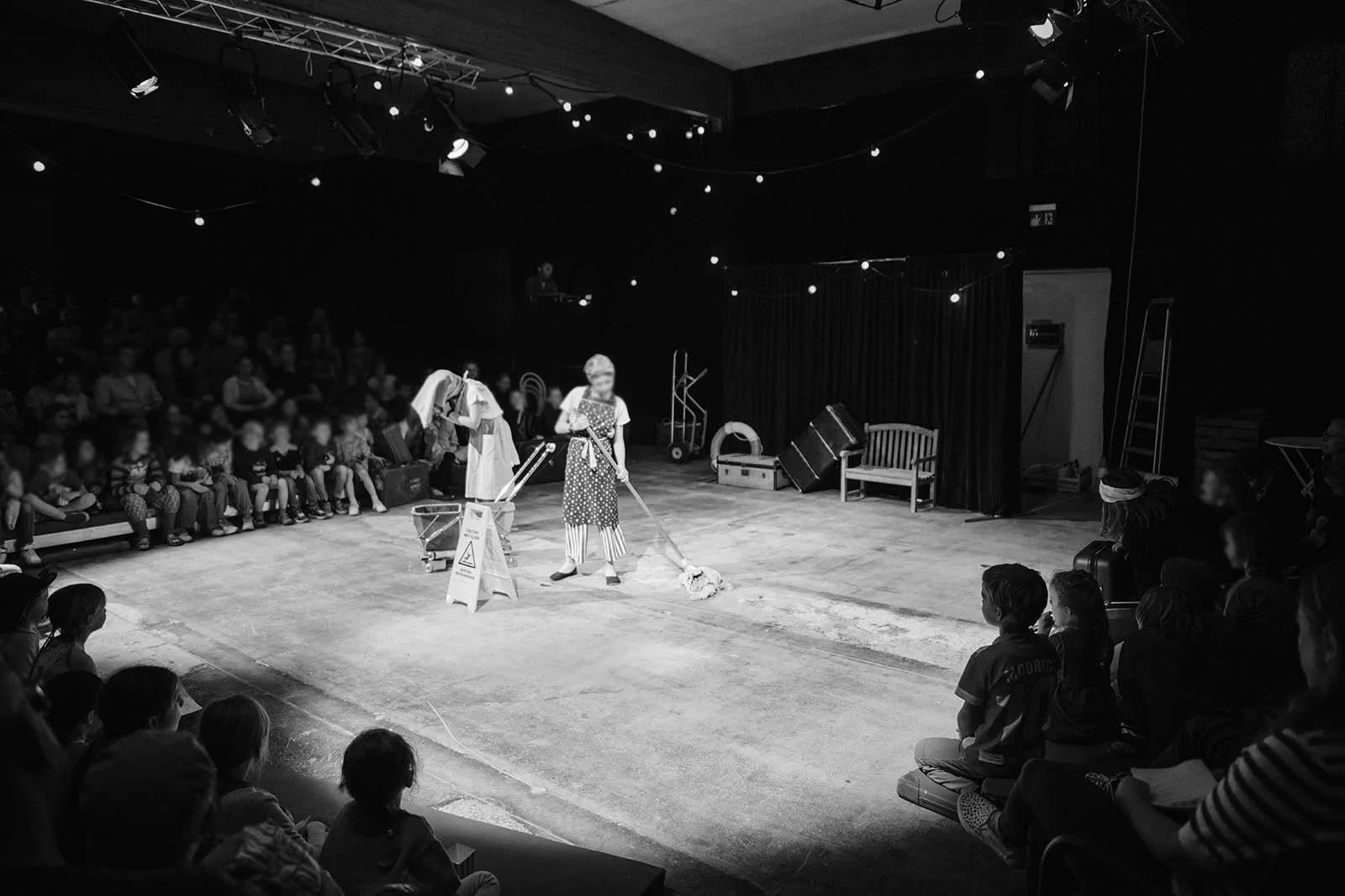 Menschen beobachten eine Theatervorstellung auf einer Bühne mit einem Clown, der den Boden reinigt, umgeben von Bühnenelementen, im Hintergrund eine schwarze Bühne mit Vorhängen und schwarzen Lichtstrahlern.