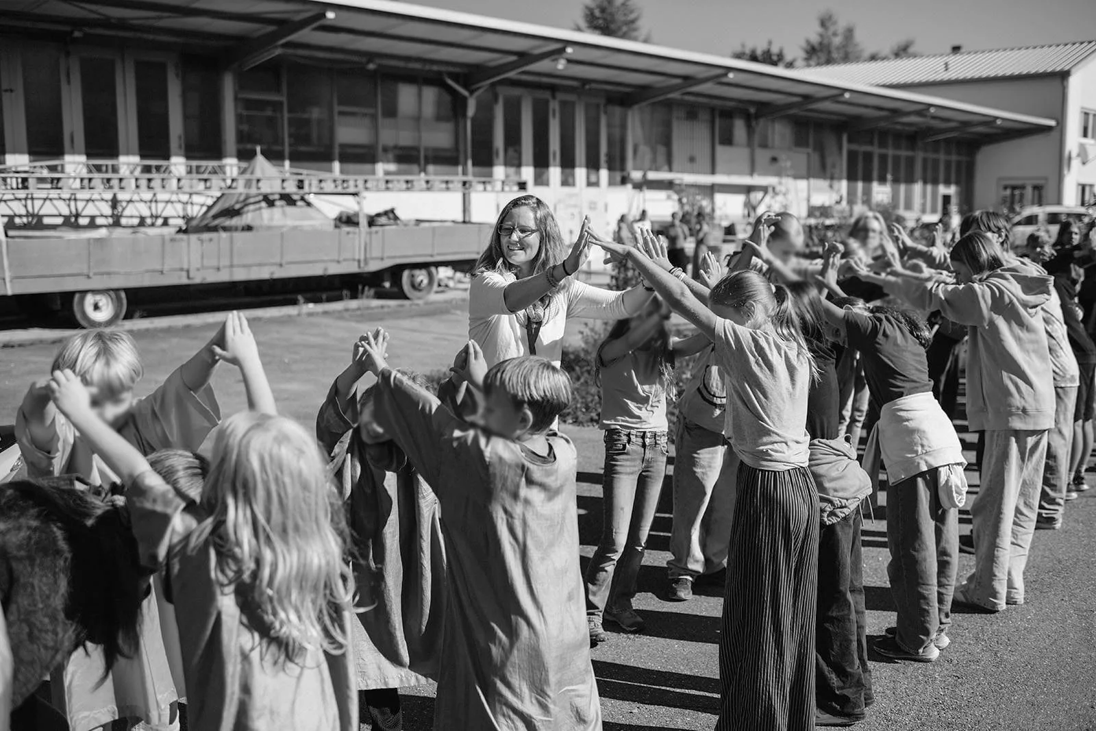 Kinder, die in einer Reihe stehen und sich gegenseitig die Hände geben, während eine erwachsene Frau ihnen dabei hilft, auf einem Schulhof oder in einem Außenbereich zu spielen.