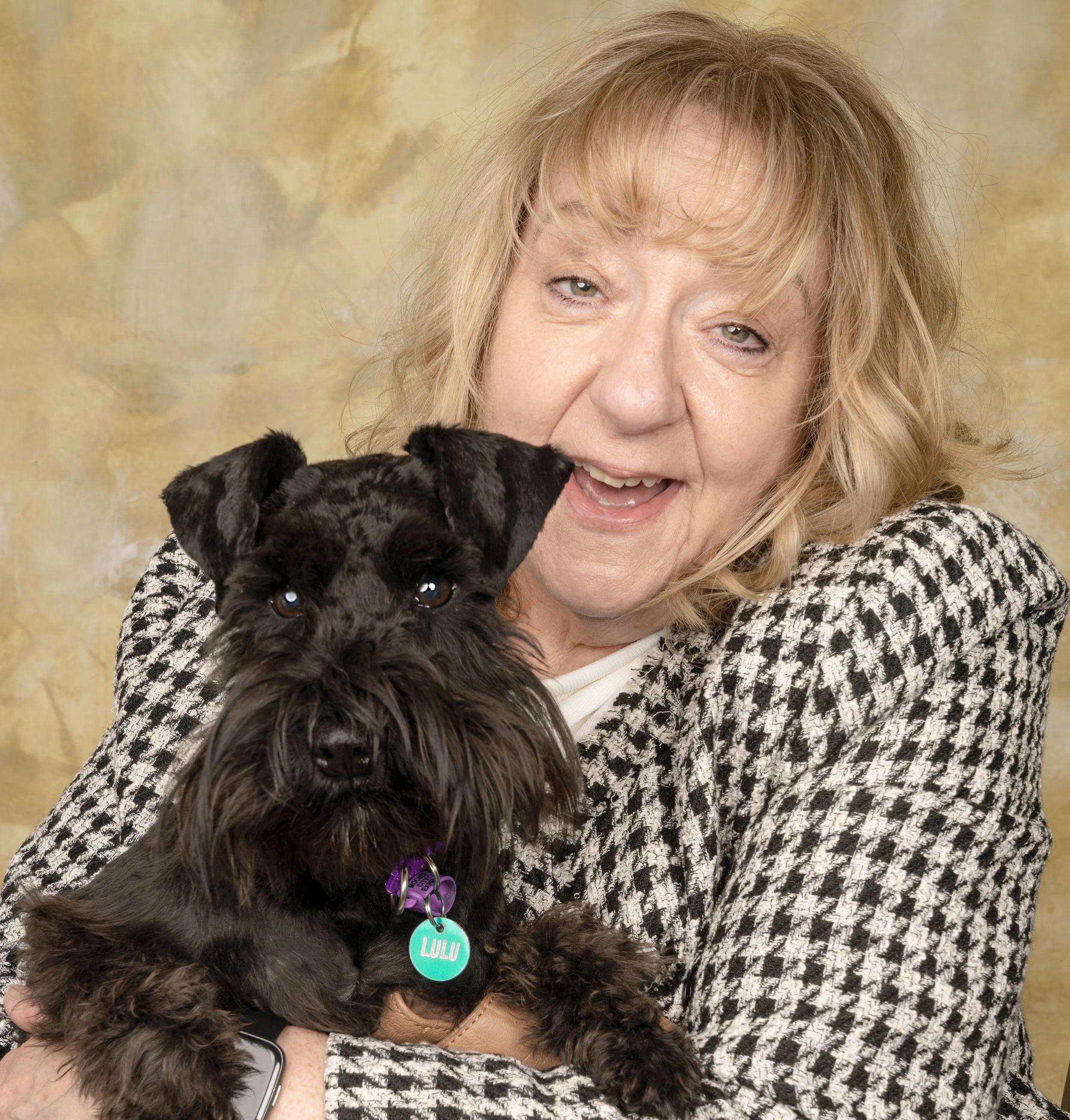 A close-up photo of a woman with light skin, blonde curly hair, and wearing a black and white patterned sweater. She is cuddling a small black puppy with dark fur, bright eyes, and a purple collar with a round tag. The background is a soft yellowish color.