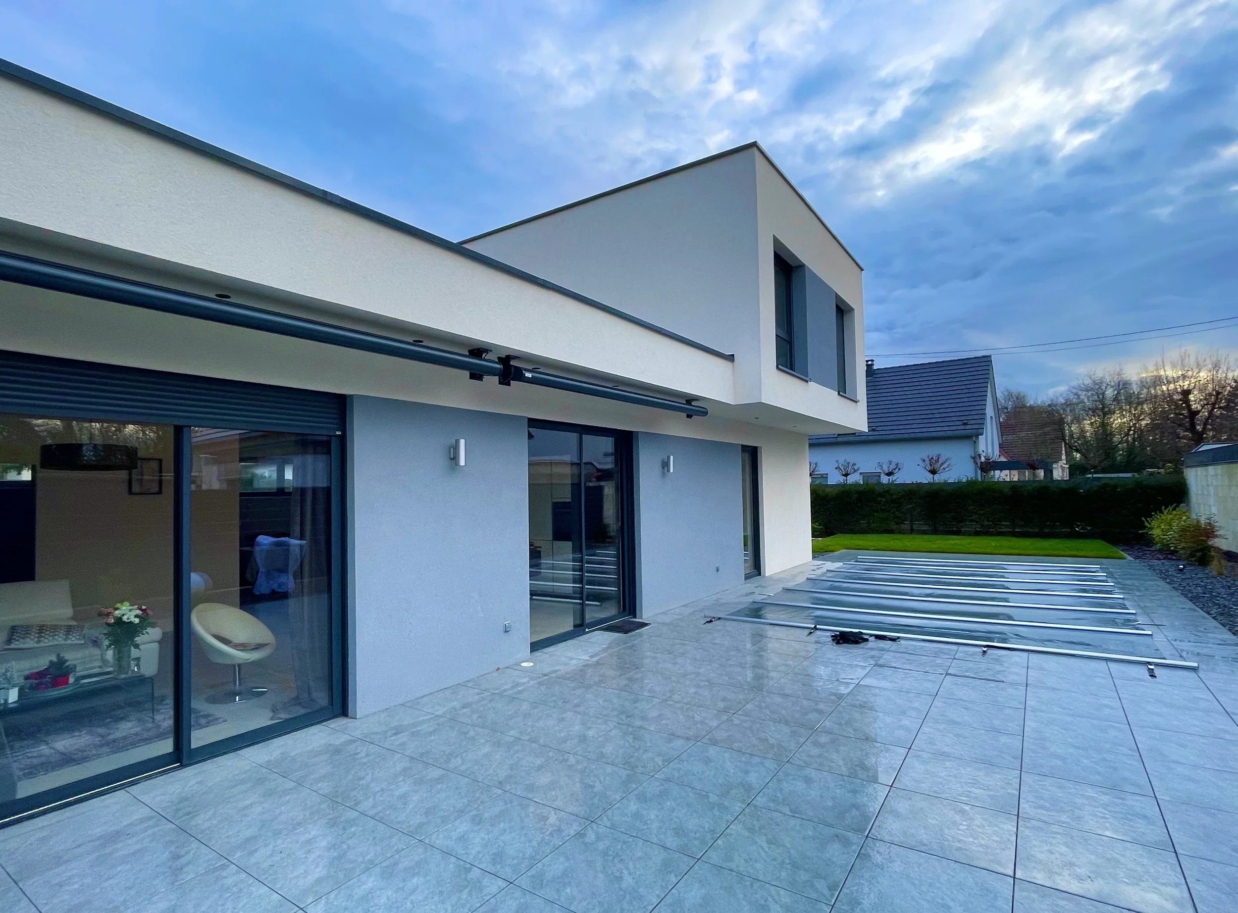 Une maison moderne avec une terrasse en pierre, de grandes fenêtres en verre, un jardin bien entretenu, et un ciel partiellement nuageux.
