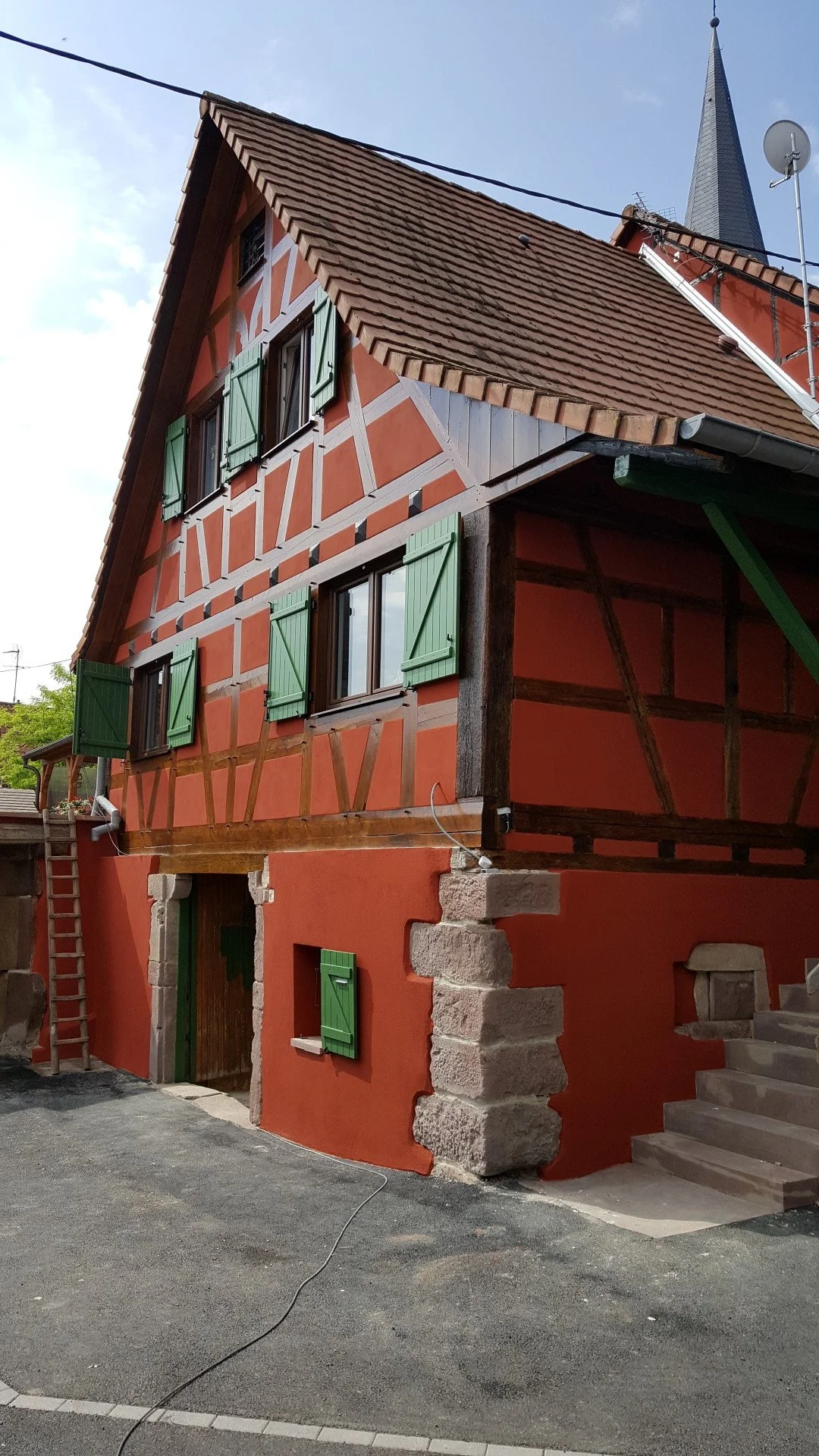 Maison à colombages avec murs en enduit orange, fenêtres à volets verts, toiture en tuiles, et une tour d'église en arrière-plan.