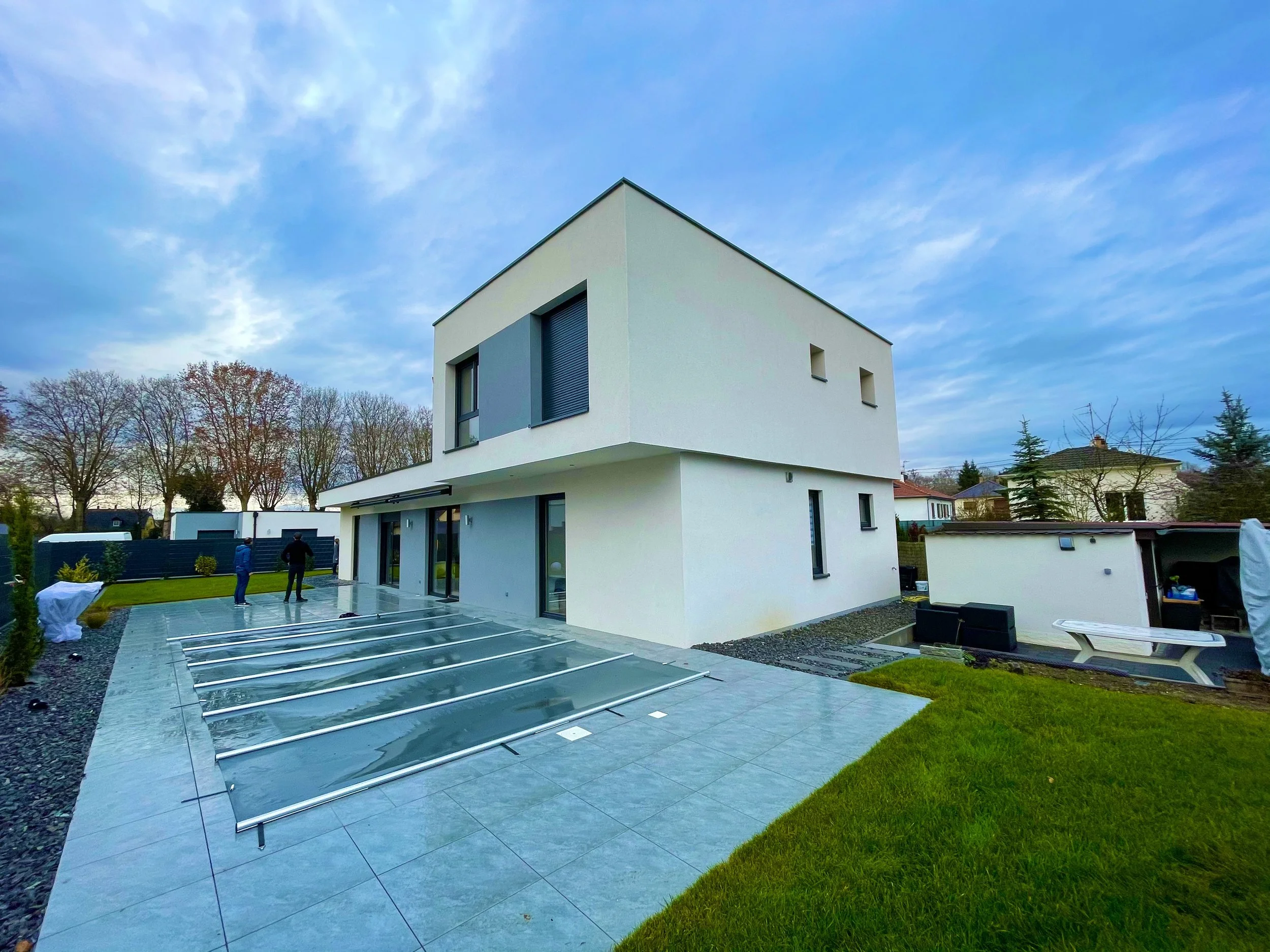 Une maison moderne blanche avec une terrasse en pierre grise, une pelouse verte, et un ciel bleu avec quelques nuages.