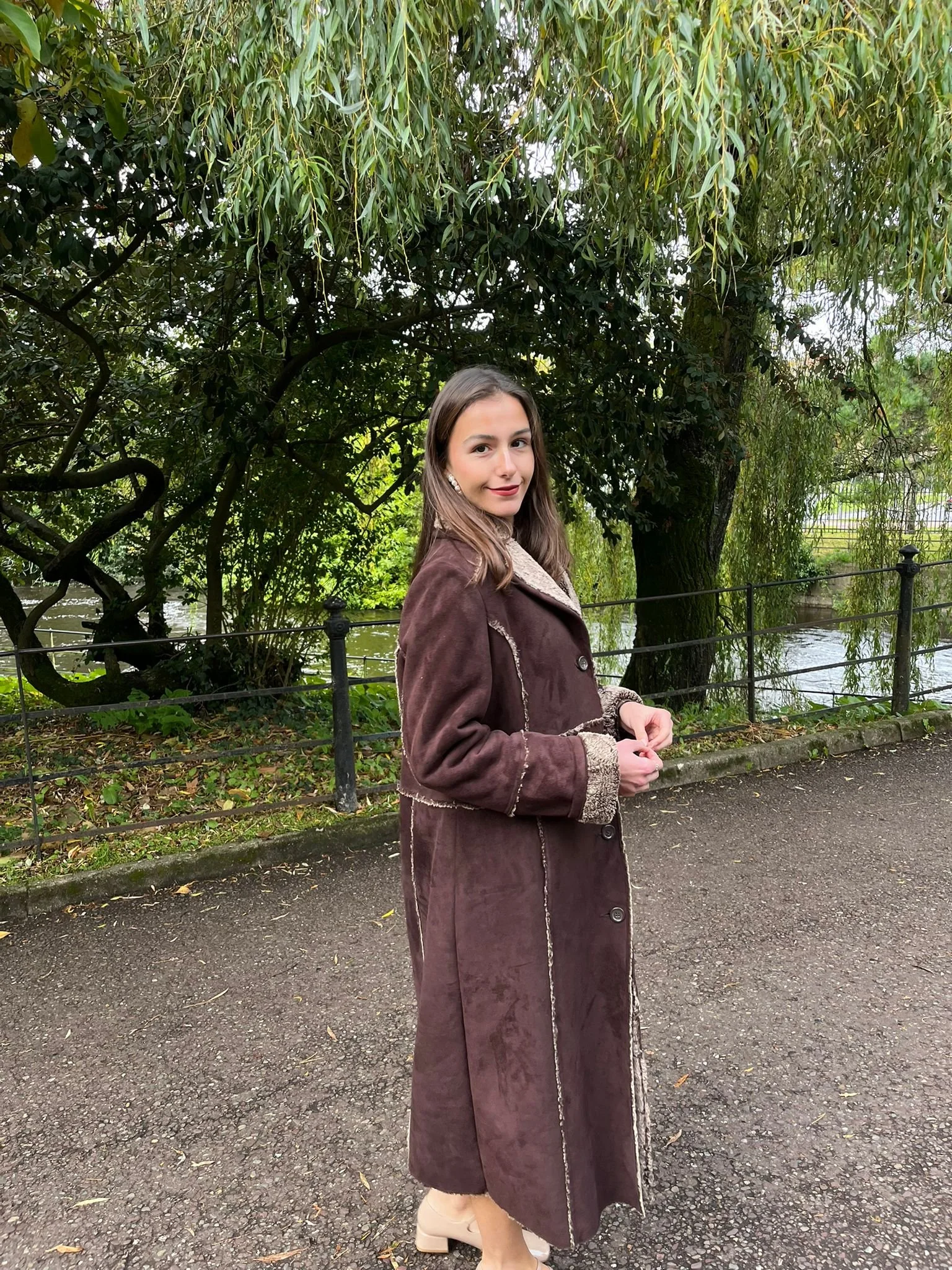 A woman with brown hair and makeup, wearing a brown long coat with a shearling collar, standing outdoors on a paved path with a metal railing, lush green trees and a river in the background.