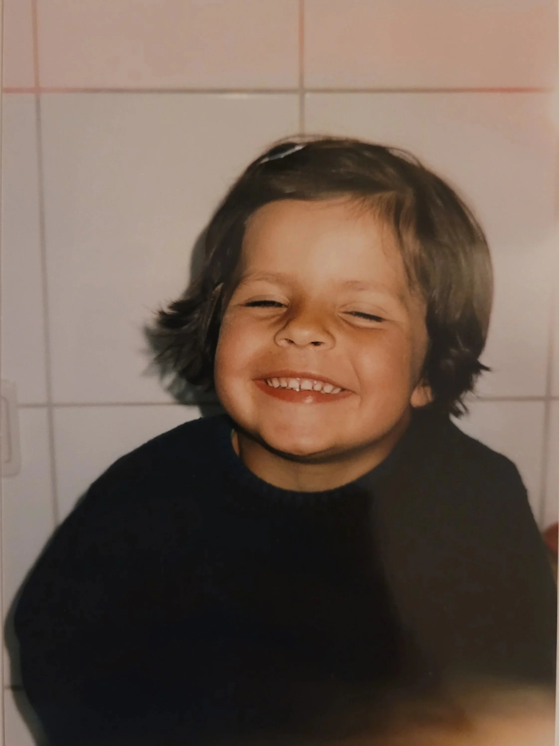 Ein lachender Junge mit dunklen Haaren vor einer weißen Fliese