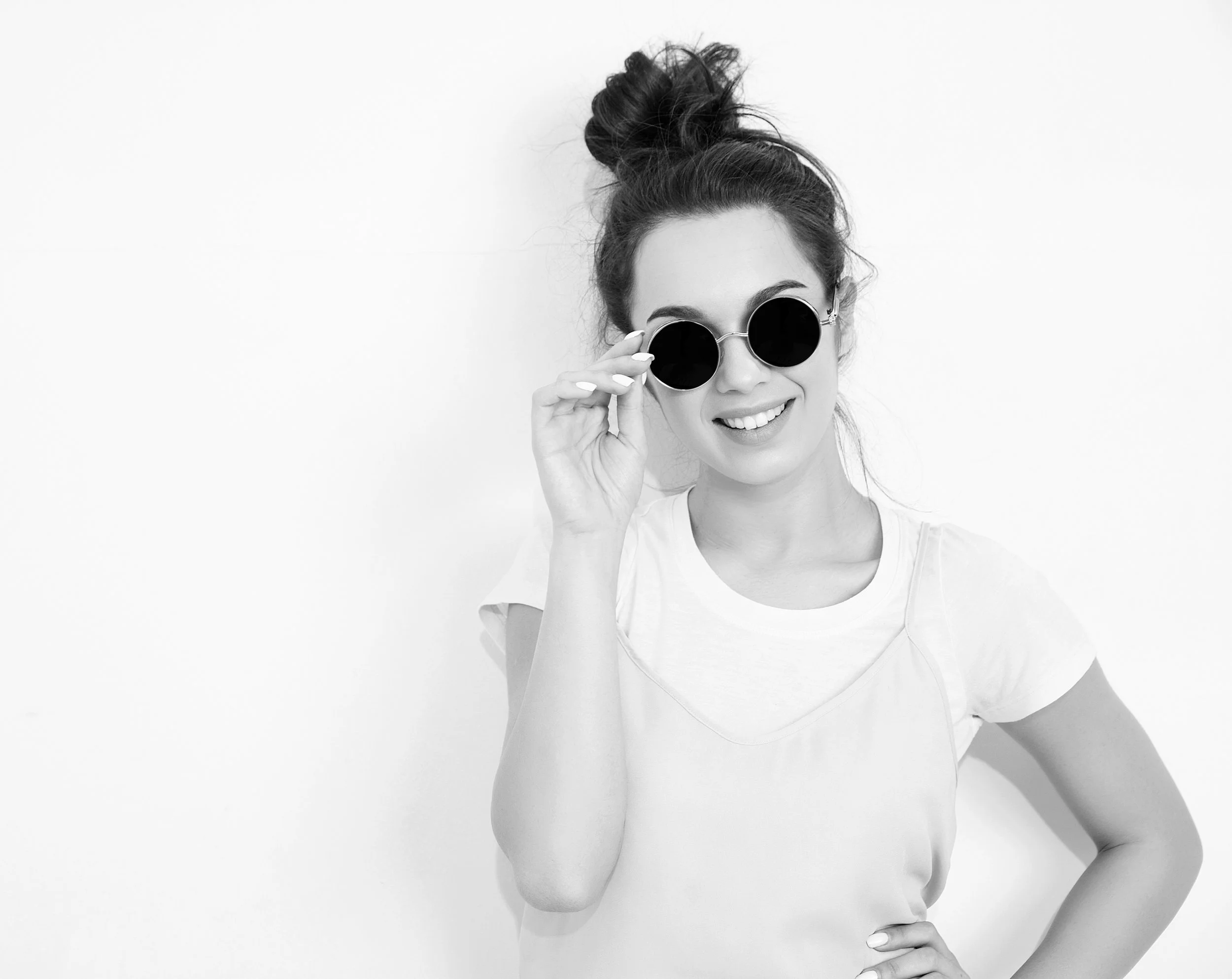 Mujer joven con peinado recogido, usando gafas de sol oscuras, sonriendo y tocándose las gafas mientras posa contra fondo blanco, vista superior de una parte del torso y la cabeza.